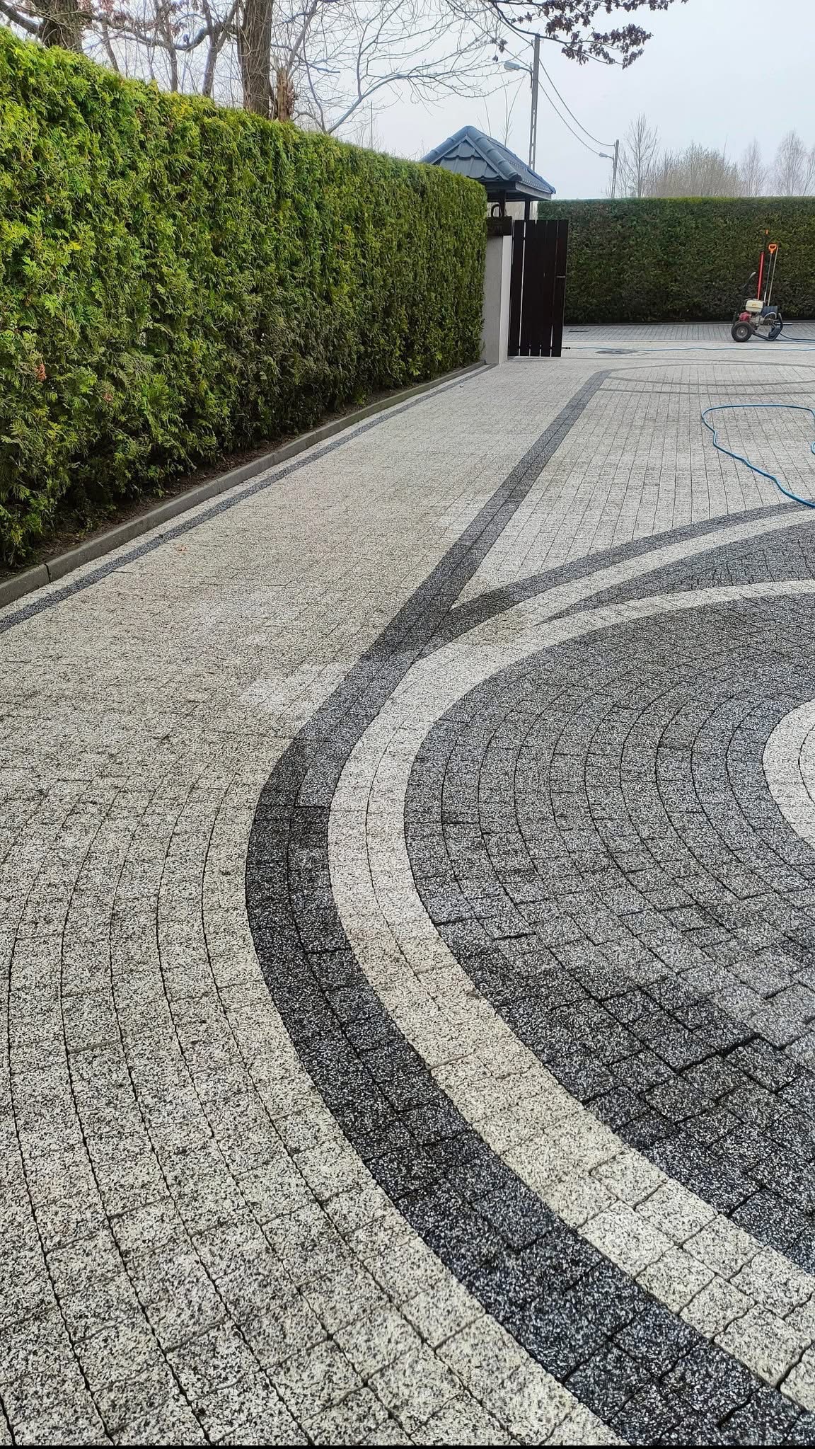 Wzorzysta kostka brukowa po umyciu, z widocznymi śladami czyszczenia. Tuje w tle, wąż do myjki ciśnieniowej i myjka ciśnieniowa po prawej stronie.