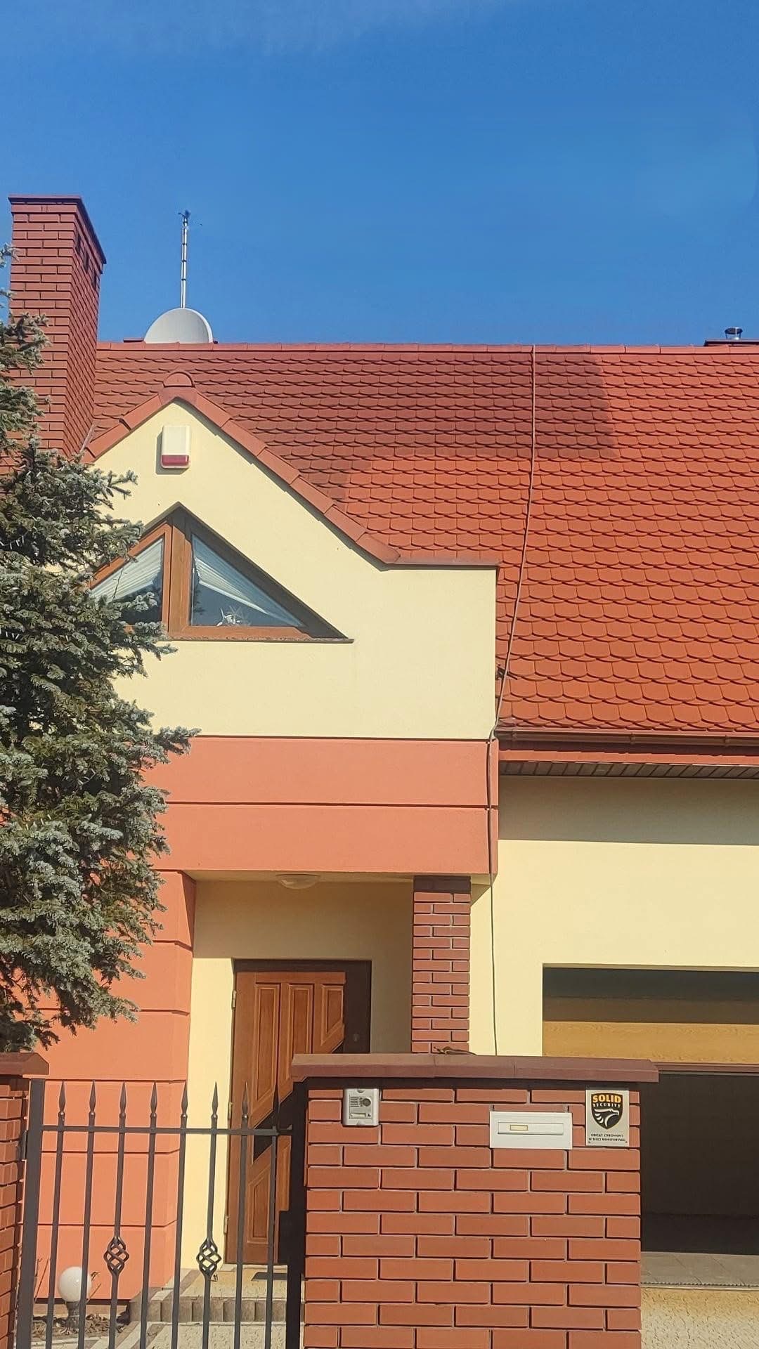 Częściowy widok domu jednorodzinnego z czerwoną dachówką, anteną satelitarną i trójkątnym oknem. Elewacja w odcieniach beżu i pomarańczy, z widocznym fragmentem ogrodzenia.