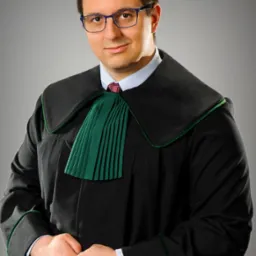 Portret adwokata z Opola w todze, z rękami splecionymi na wysokości pasa. Mężczyzna w okularach, z subtelnym uśmiechem, na neutralnym, szarym tle.