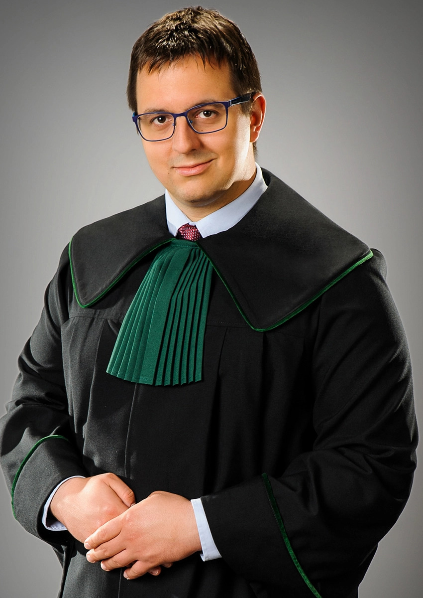 Portret adwokata z Opola w todze, z rękami splecionymi na wysokości pasa. Mężczyzna w okularach, z subtelnym uśmiechem, na neutralnym, szarym tle.