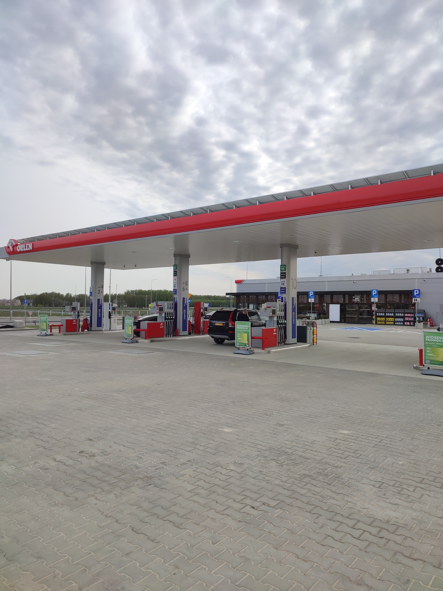 Stacja benzynowa Orlen z dystrybutorami paliw, samochodem i budynkiem sklepu w tle. Widok na pusty plac z szarej kostki brukowej i pochmurne niebo.