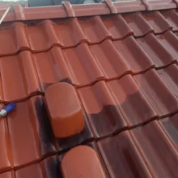 Częściowo umyty, błyszczący dach z czerwonej dachówki ceramicznej. Widoczna różnica w kolorze między czystą a brudną powierzchnią. W tle fragment budynku.