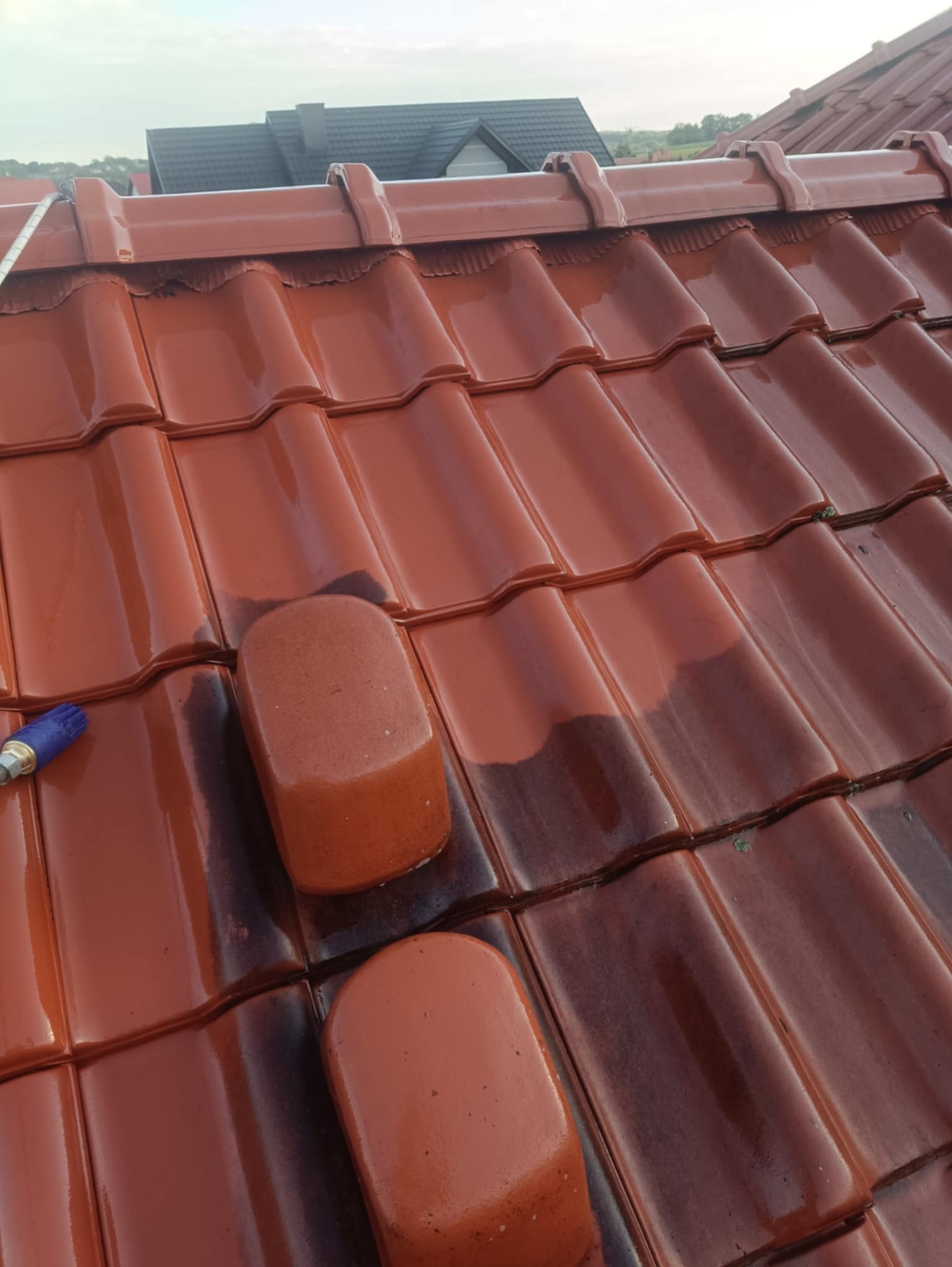 Częściowo umyty, błyszczący dach z czerwonej dachówki ceramicznej. Widoczna różnica w kolorze między czystą a brudną powierzchnią. W tle fragment budynku.