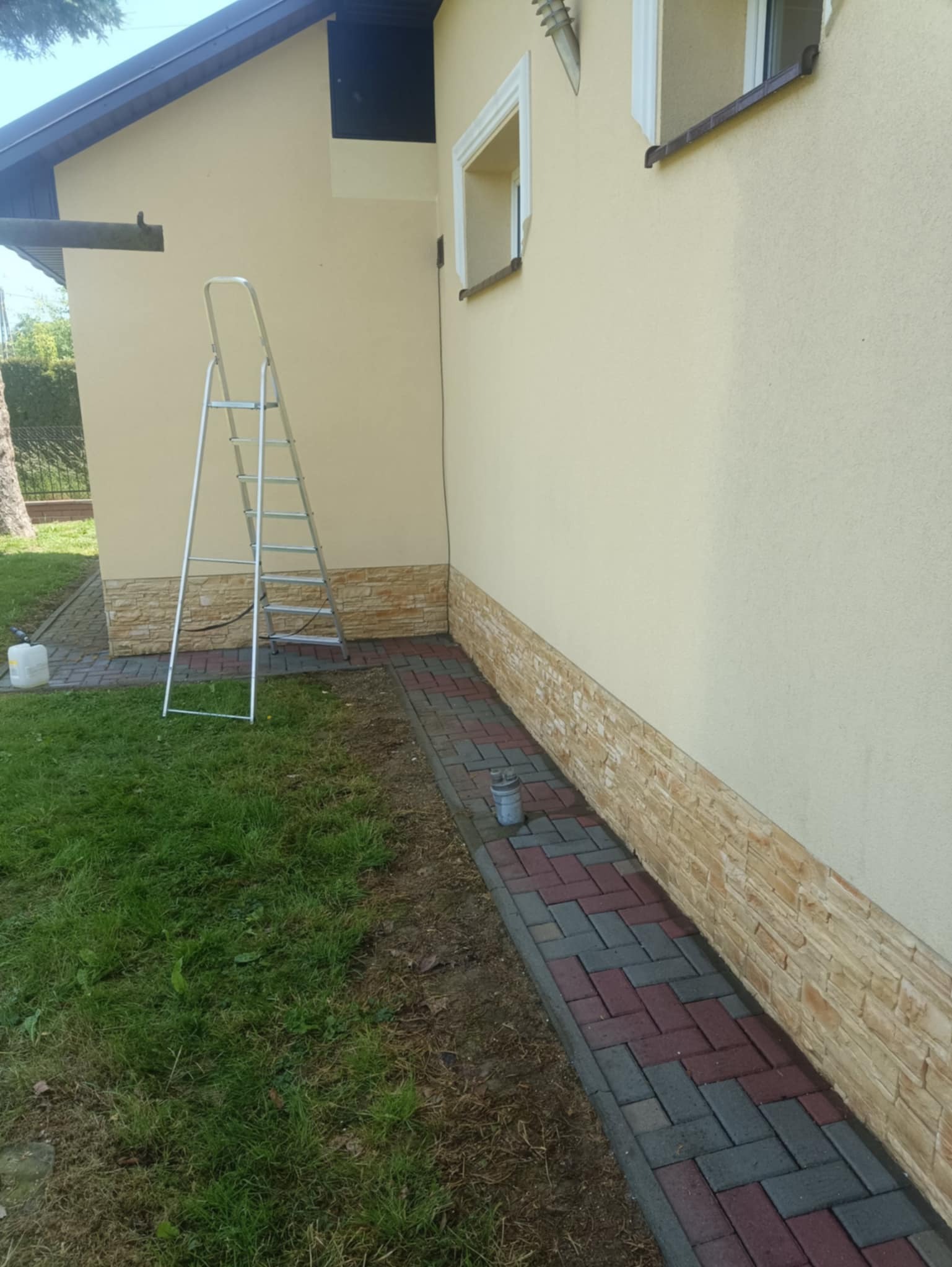Elewacja domu z dekoracyjnym kamieniem i kostką brukową, drabina oparta o ścianę, przygotowania do prac na zewnątrz budynku.