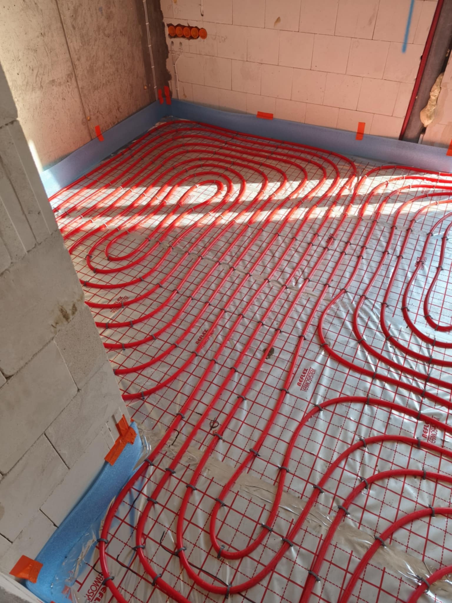 Czerwone rury ułożone na izolacji, przygotowane do zalania posadzką w systemie ogrzewania. Widok na instalację w surowym wnętrzu.