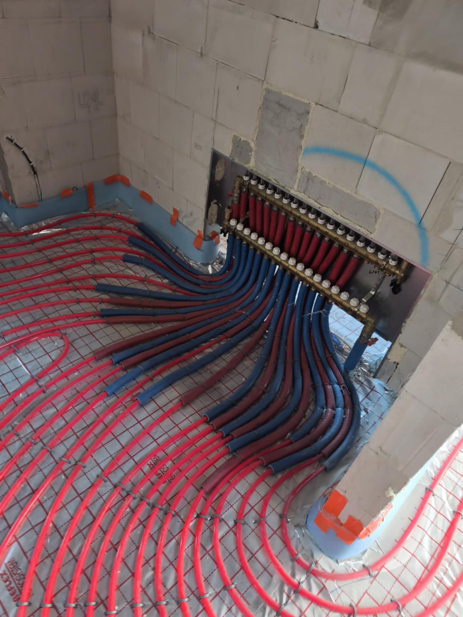 Instalacja ogrzewania podłogowego w surowym wnętrzu: czerwone rury na folii, niebieskie rury doprowadzające do rozdzielacza. Widoczne ściany z betonu komórkowego.