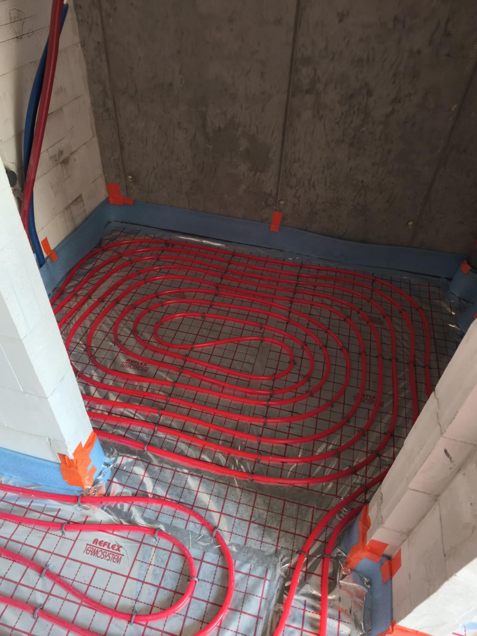 Czerwone rury systemu ogrzewania podłogowego ułożone na folii z siatką, przymocowane do ścian z bloczków. Widoczne elementy montażowe.