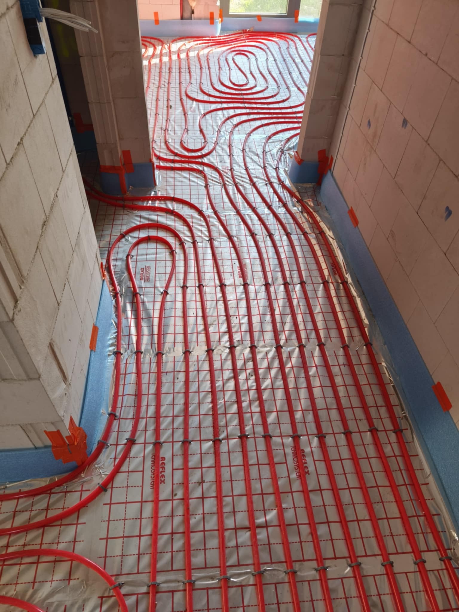 Czerwone rury systemu rozprowadzone na izolacji podłogowej z siatką, przygotowane do zalania wylewką. Widok z góry na etapie montażu w nowym budynku.