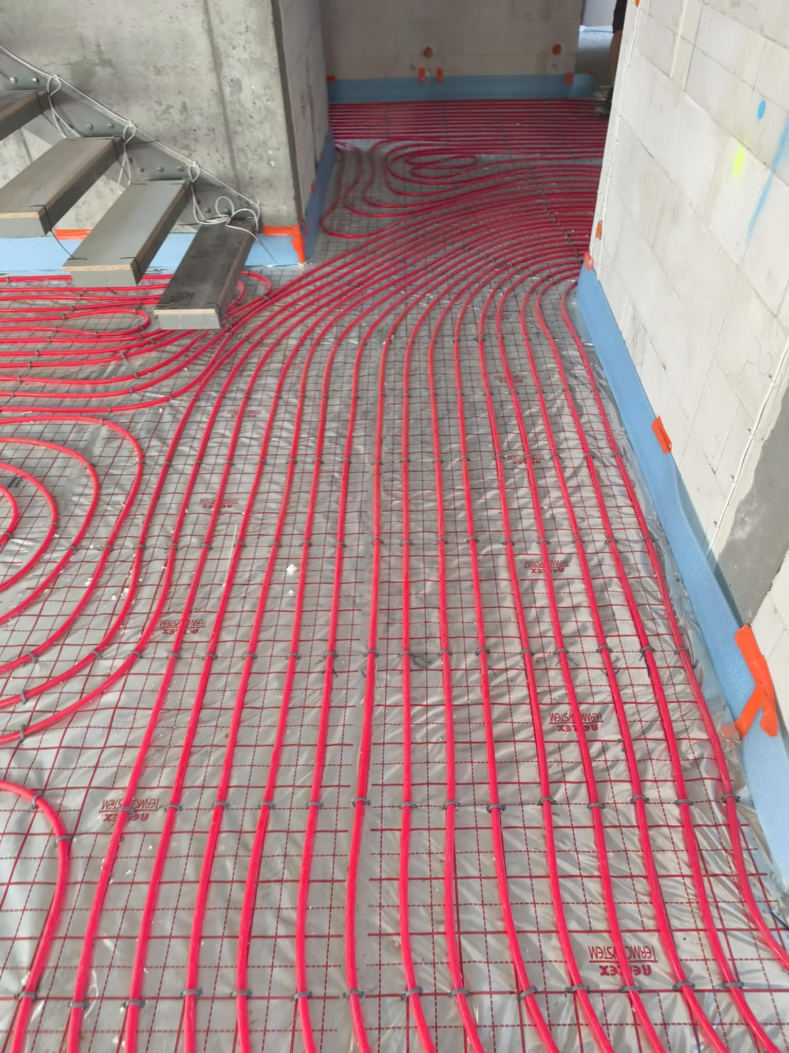 Czerwone rury ogrzewania na macie izolacyjnej, widok z góry, instalacja w trakcie montażu przy schodach, przygotowanie do wylewki.