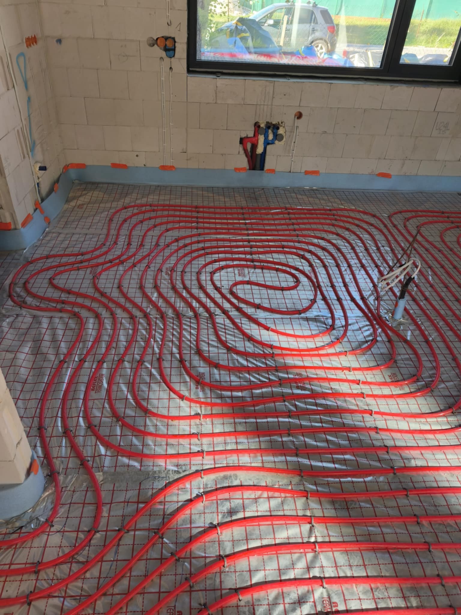 Czerwone rury ułożone spiralnie na szarej macie w trakcie montażu. Widoczne przyłącza do instalacji CO i CWU na ścianie. Budynek w trakcie budowy.