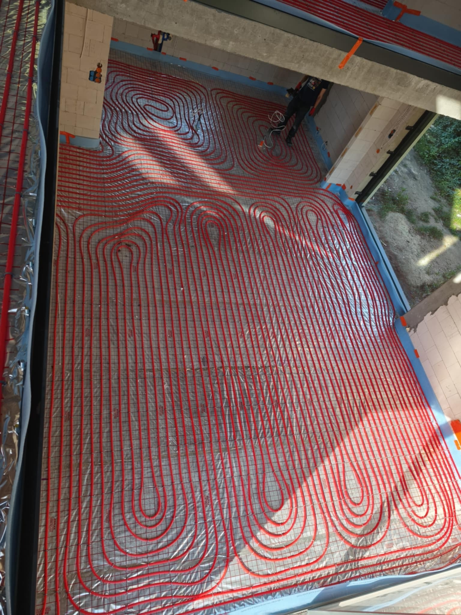 Czerwone rury ułożone na folii aluminiowej, tworzące pętle ogrzewania podłogowego w surowym wnętrzu budynku. Widoczny instalator przy pracy.