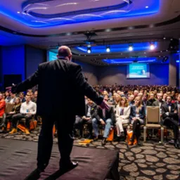 Prelegent w garniturze przemawia do dużej publiczności w sali konferencyjnej oświetlonej niebieskim światłem, z widocznym ekranem projekcyjnym w tle.