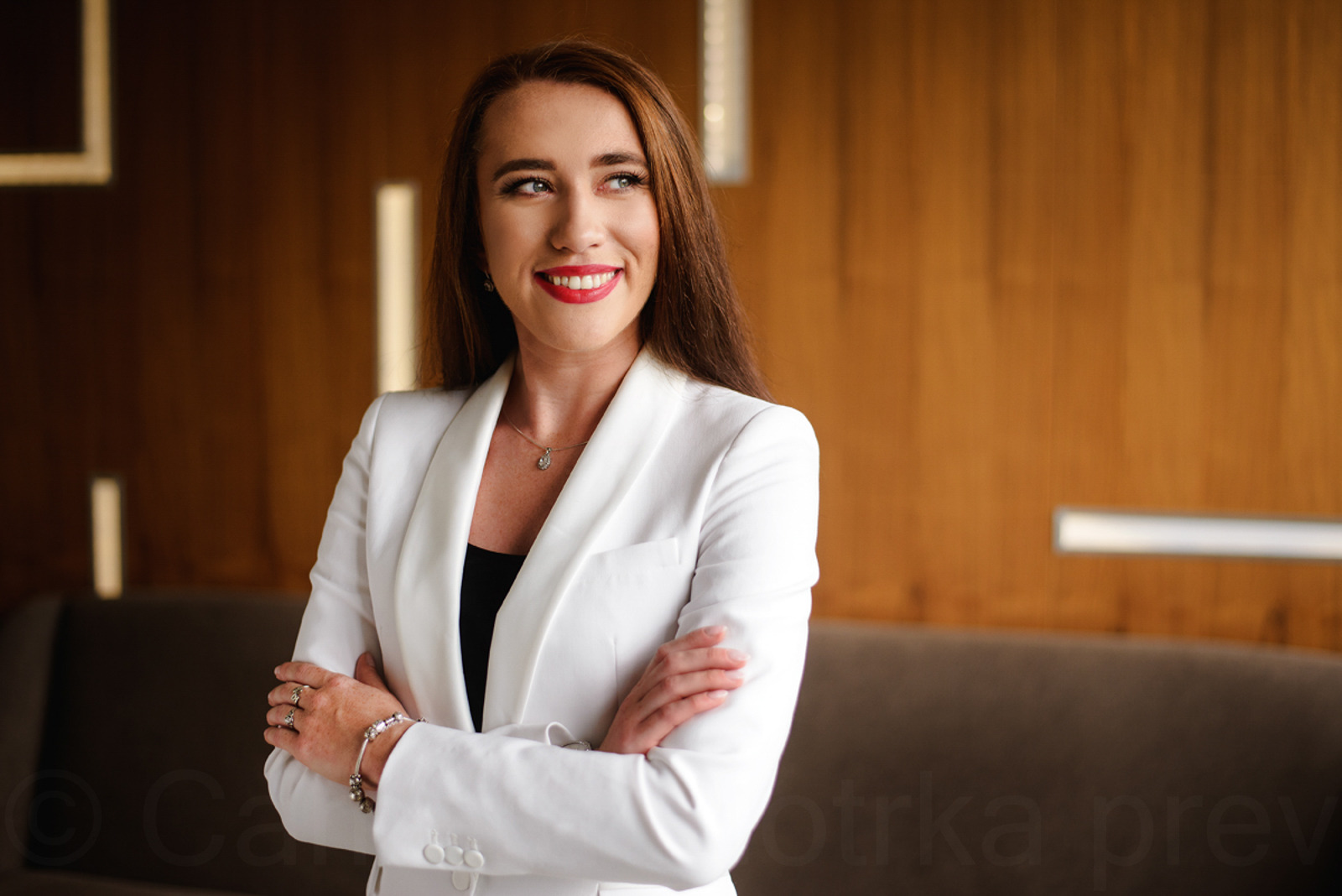 Portret uśmiechniętej kobiety w białym żakiecie, skrzyżowane ręce, elegancka biżuteria, stonowane tło z drewnianą ścianą. Sesja biznesowa w studio.