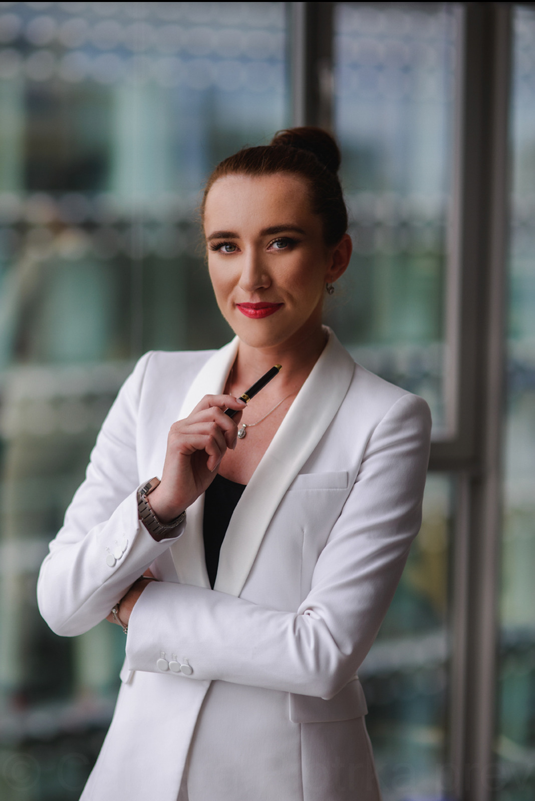 Portret kobiety w białym garniturze, z czarnym długopisem w dłoni, na tle przeszklonego biura. Elegancki strój, profesjonalny wizerunek.