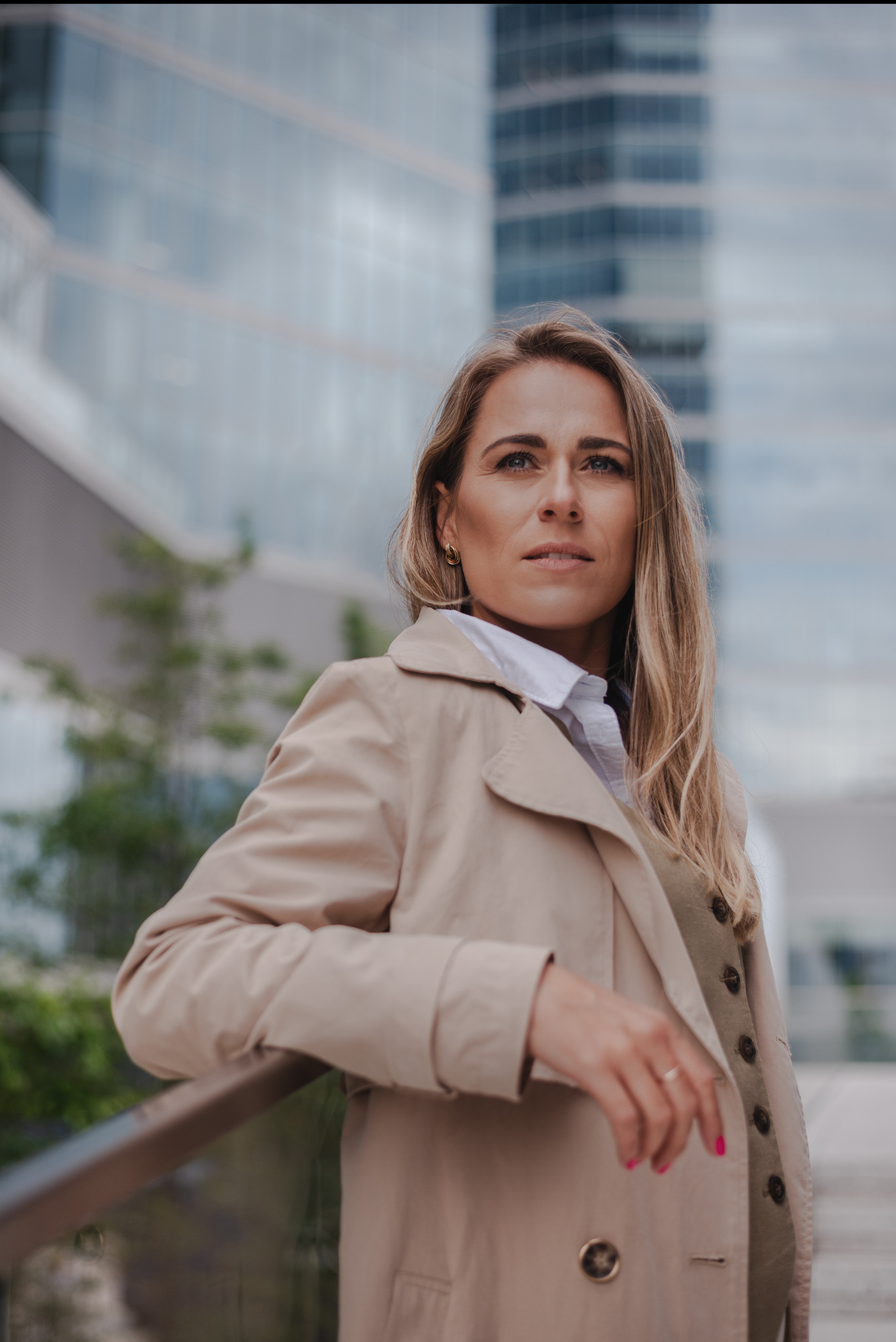Portret kobiety w beżowym płaszczu na tle nowoczesnego biurowca. Uwagę przyciągają złote kolczyki i różowy manicure.