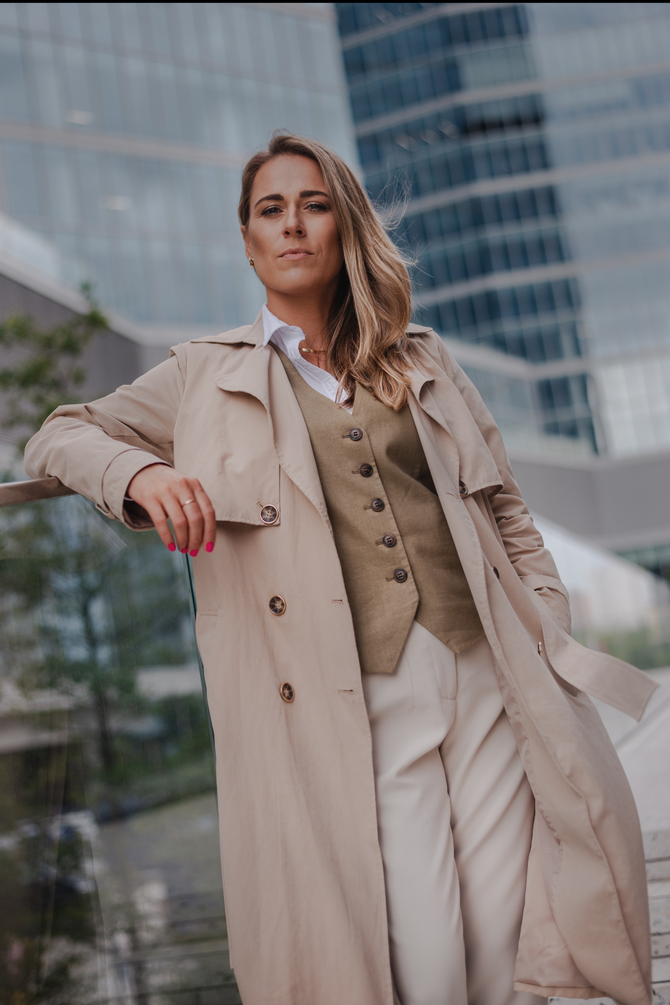Profesjonalna sesja w Górze Kalwarii: Kobieta w beżowym płaszczu i kamizelce pozuje na tle nowoczesnego biurowca. Uwagę zwracają różowe paznokcie i pewny wyraz twarzy.