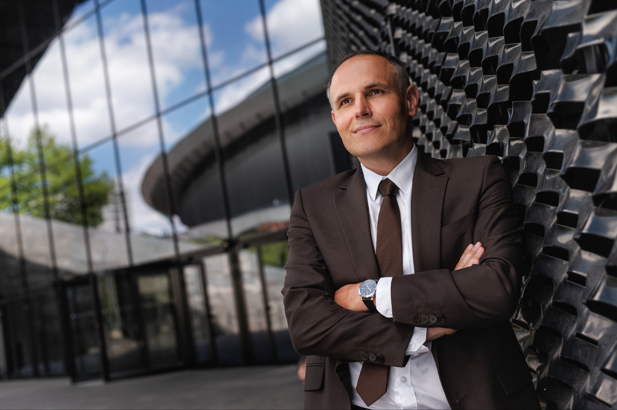 Mężczyzna w brązowym garniturze, z zegarkiem na ręku, oparty o nowoczesną, czarną ścianę na tle przeszklonego budynku. Uśmiechnięty, patrzy w górę.