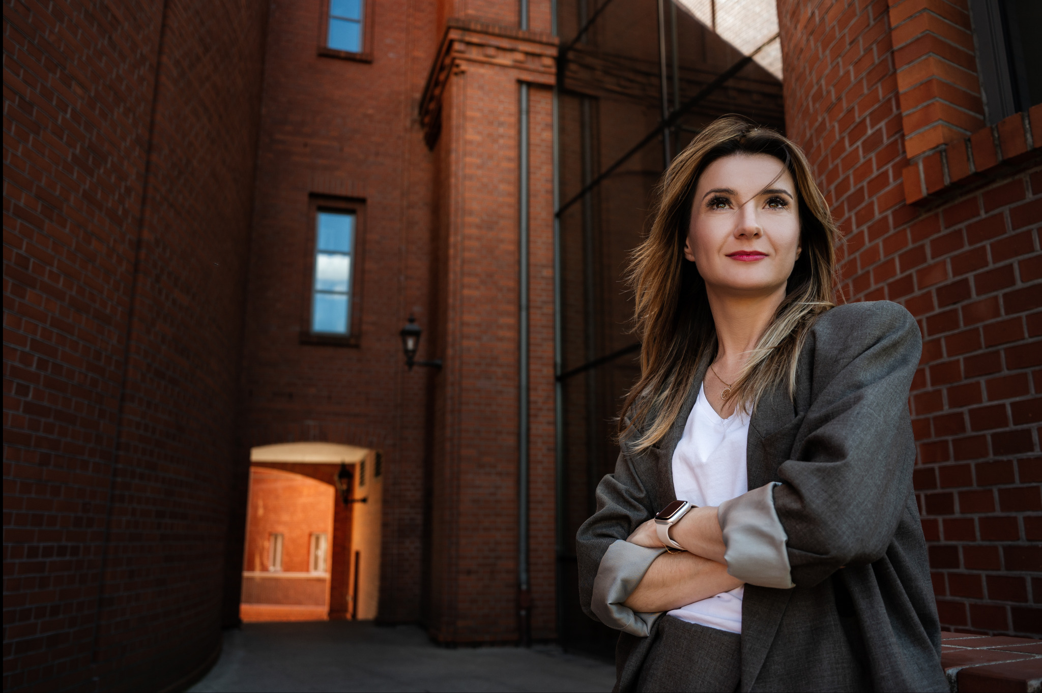 Profesjonalny portret kobiety w szarym garniturze na tle ceglanej architektury miejskiej. Ujęcie z dołu, podkreślające pewność siebie i nowoczesny styl.