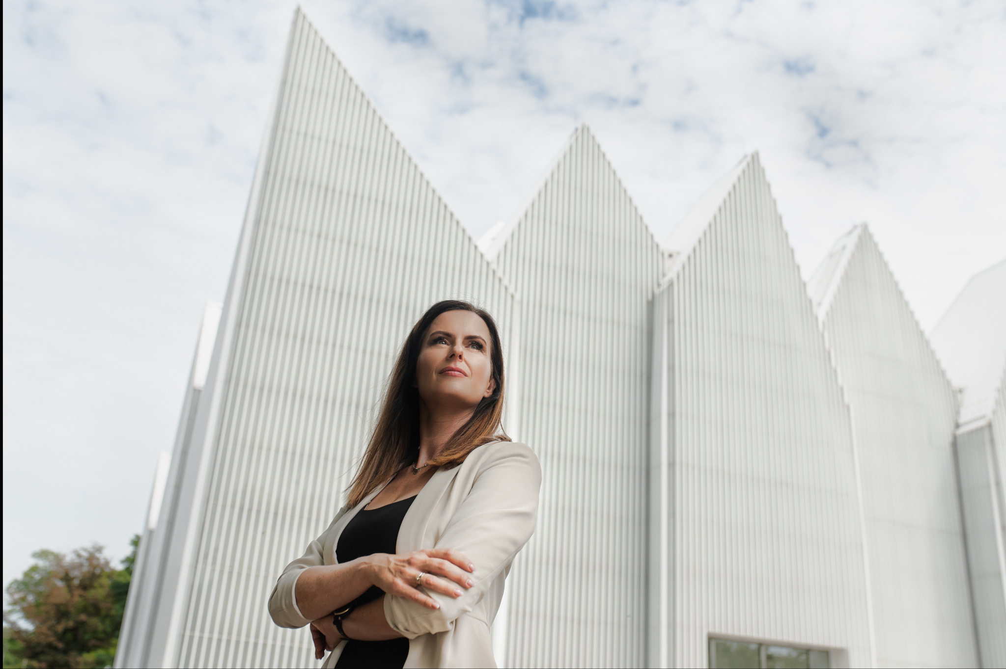 Profesjonalna sesja biznesowa: Kobieta w jasnym żakiecie, skrzyżowane ręce, patrzy w górę na tle nowoczesnej architektury i pochmurnego nieba.