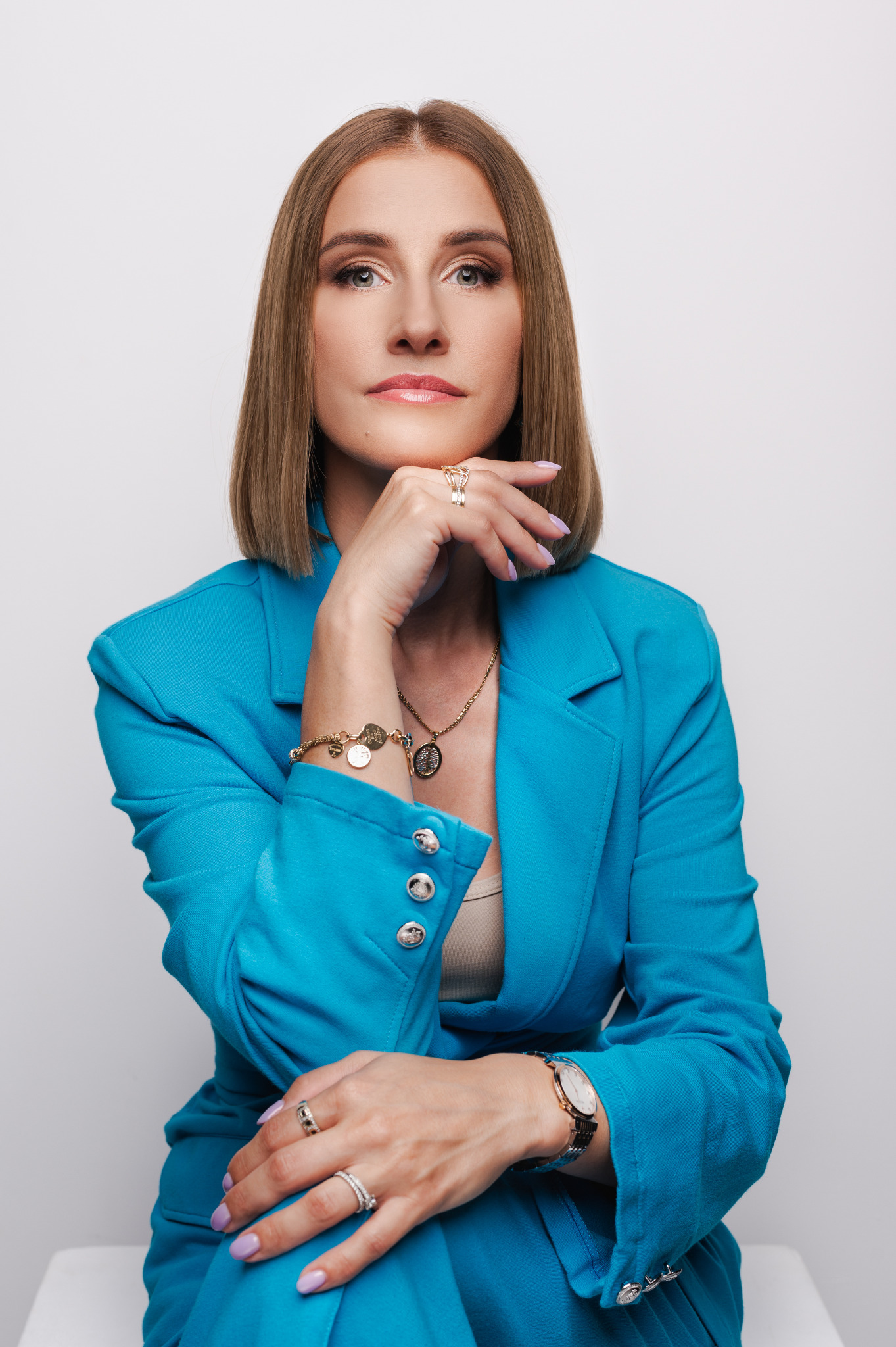 Portret kobiety w turkusowym garniturze, z biżuterią, opierającej podbródek na dłoni. Profesjonalny wizerunek na neutralnym tle, idealny do sesji biznesowej.