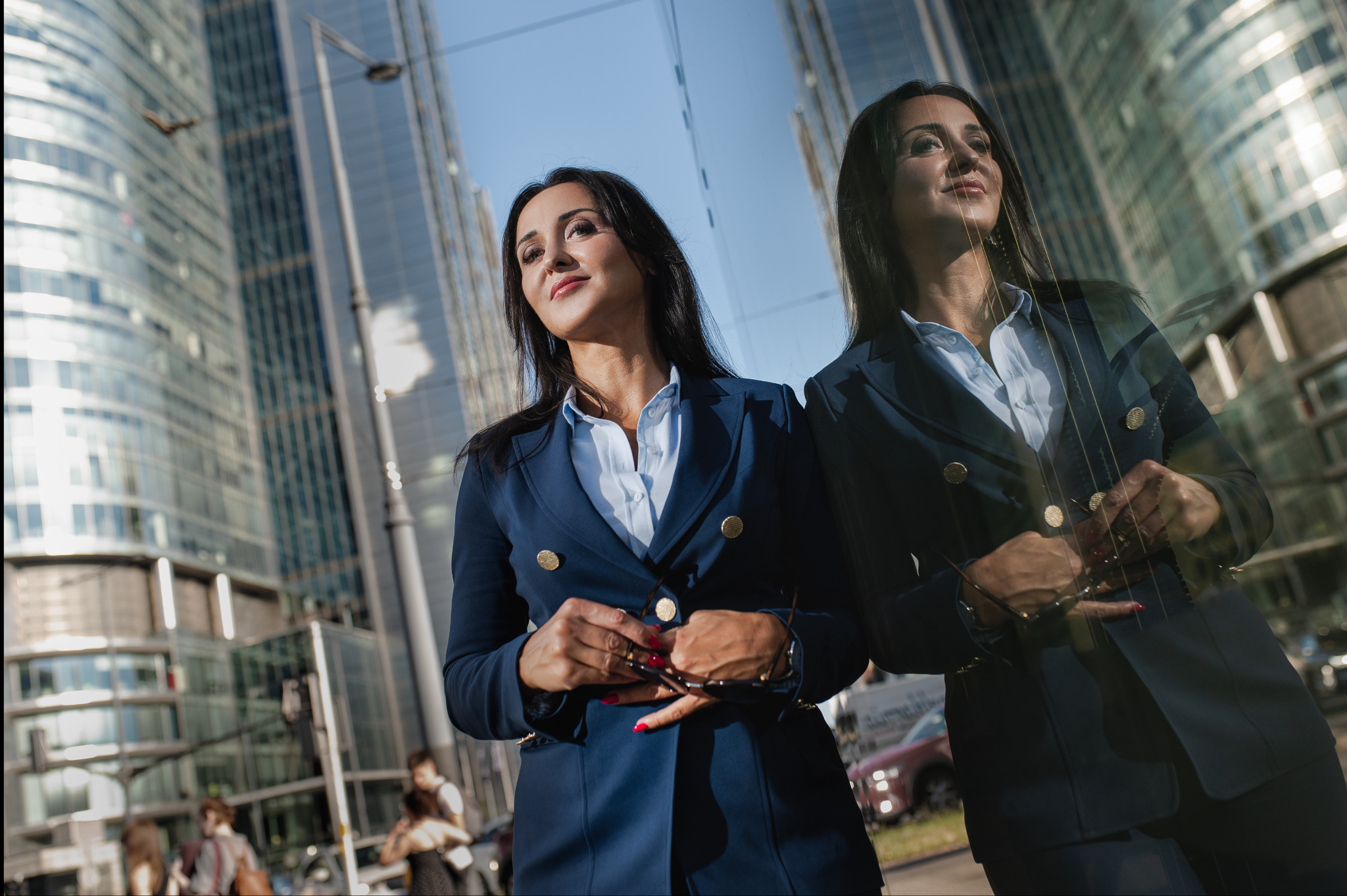 Portret kobiety w granatowym garniturze na tle biurowców, z odbiciem w szklanej fasadzie. Kadr z dołu, podkreślający profesjonalny wizerunek.