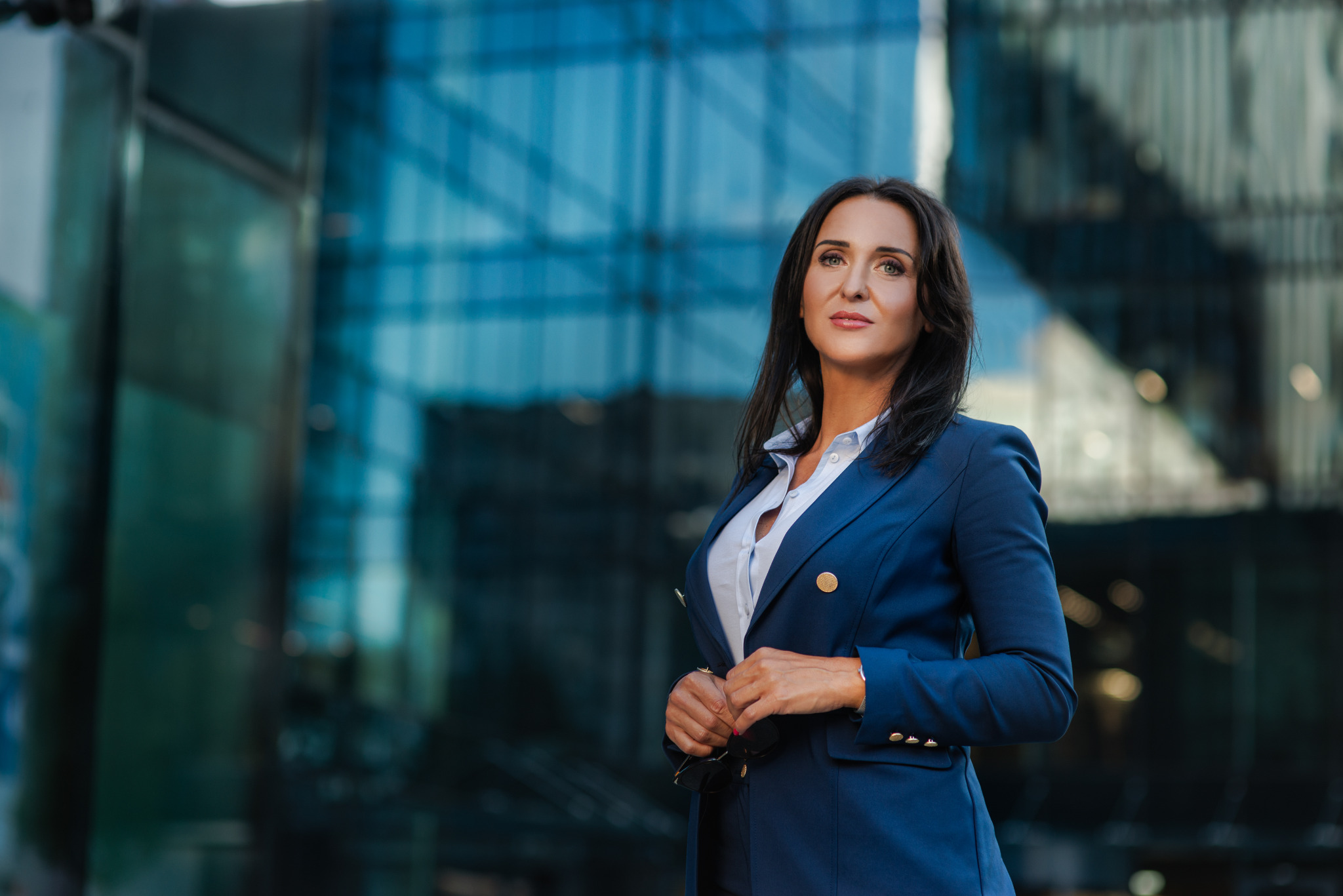 Profesjonalny portret kobiety w granatowym garniturze na tle nowoczesnego, szklanego budynku. Elegancki wizerunek biznesowy z okularami w dłoni.
