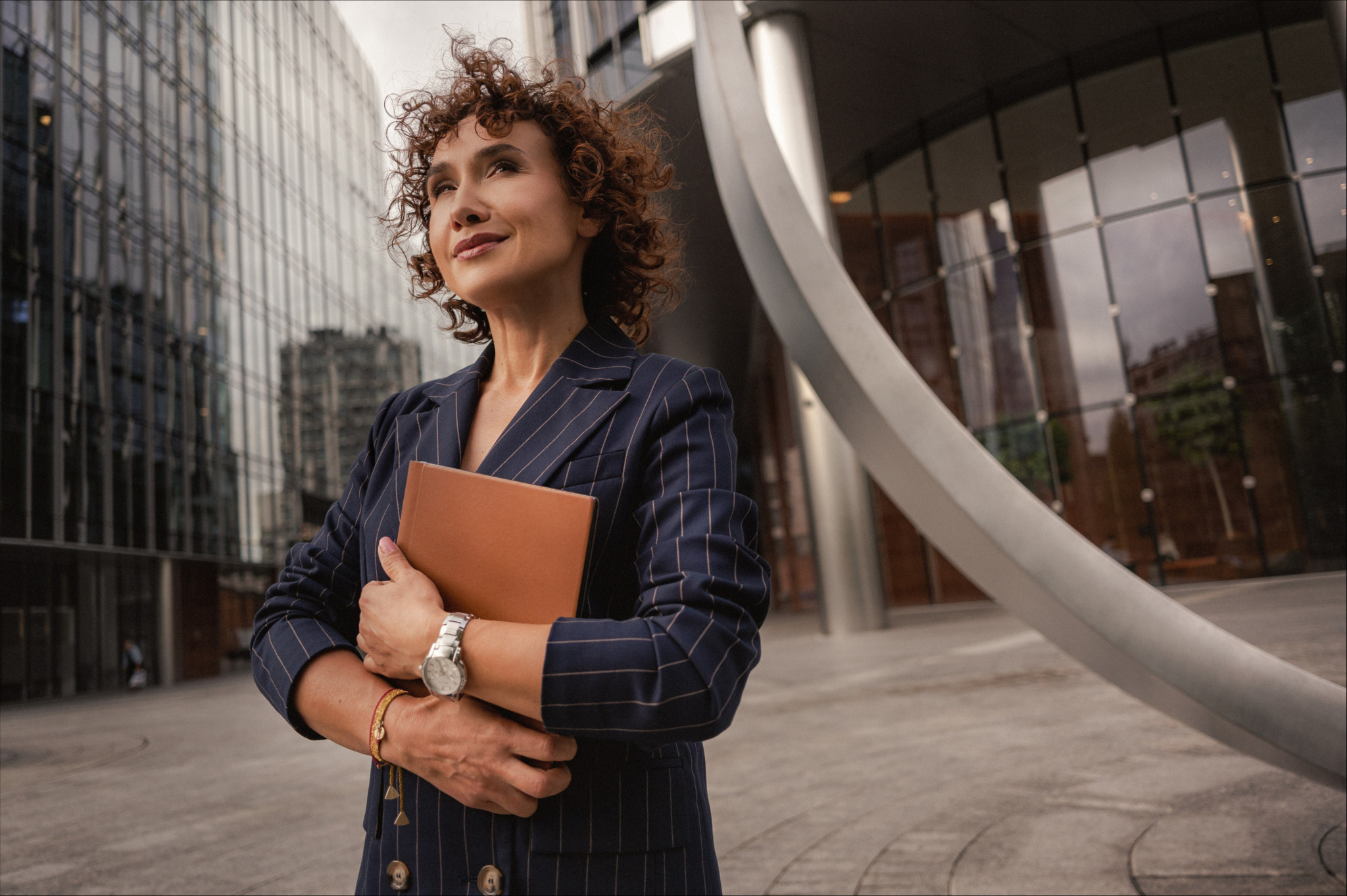 Profesjonalny portret kobiety w garniturze w paski, trzymającej brązowy notatnik na tle nowoczesnej architektury miejskiej. Spojrzenie w górę, podkreślone kręcone włosy.