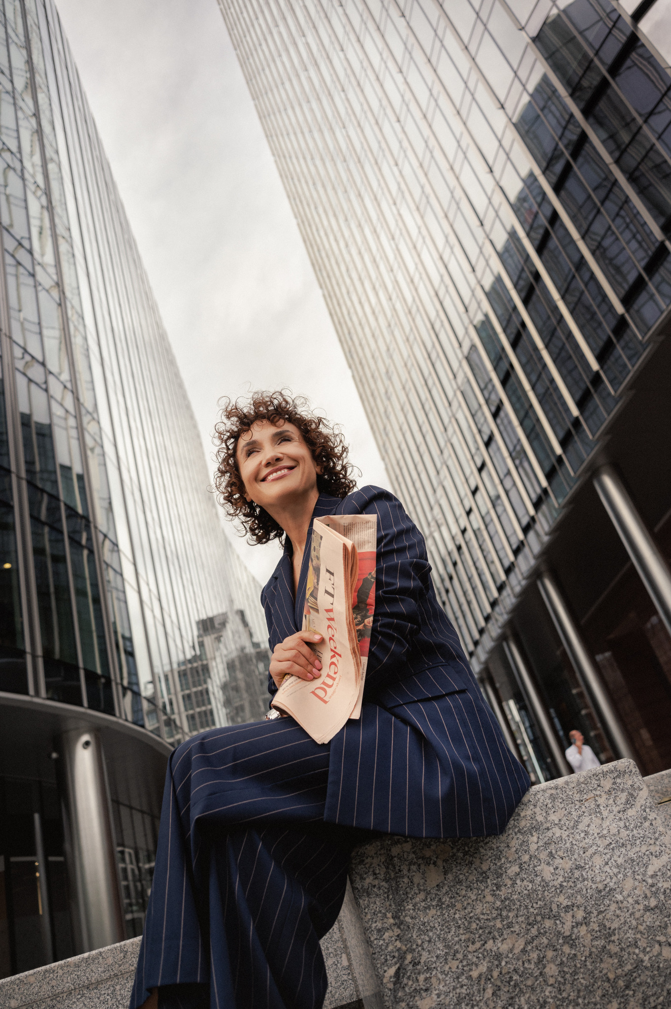 Uśmiechnięta kobieta w garniturze w paski, siedząca na tle szklanych biurowców i trzymająca gazetę 'FT Weekend'. Kadr z dołu, biznesowa elegancja.