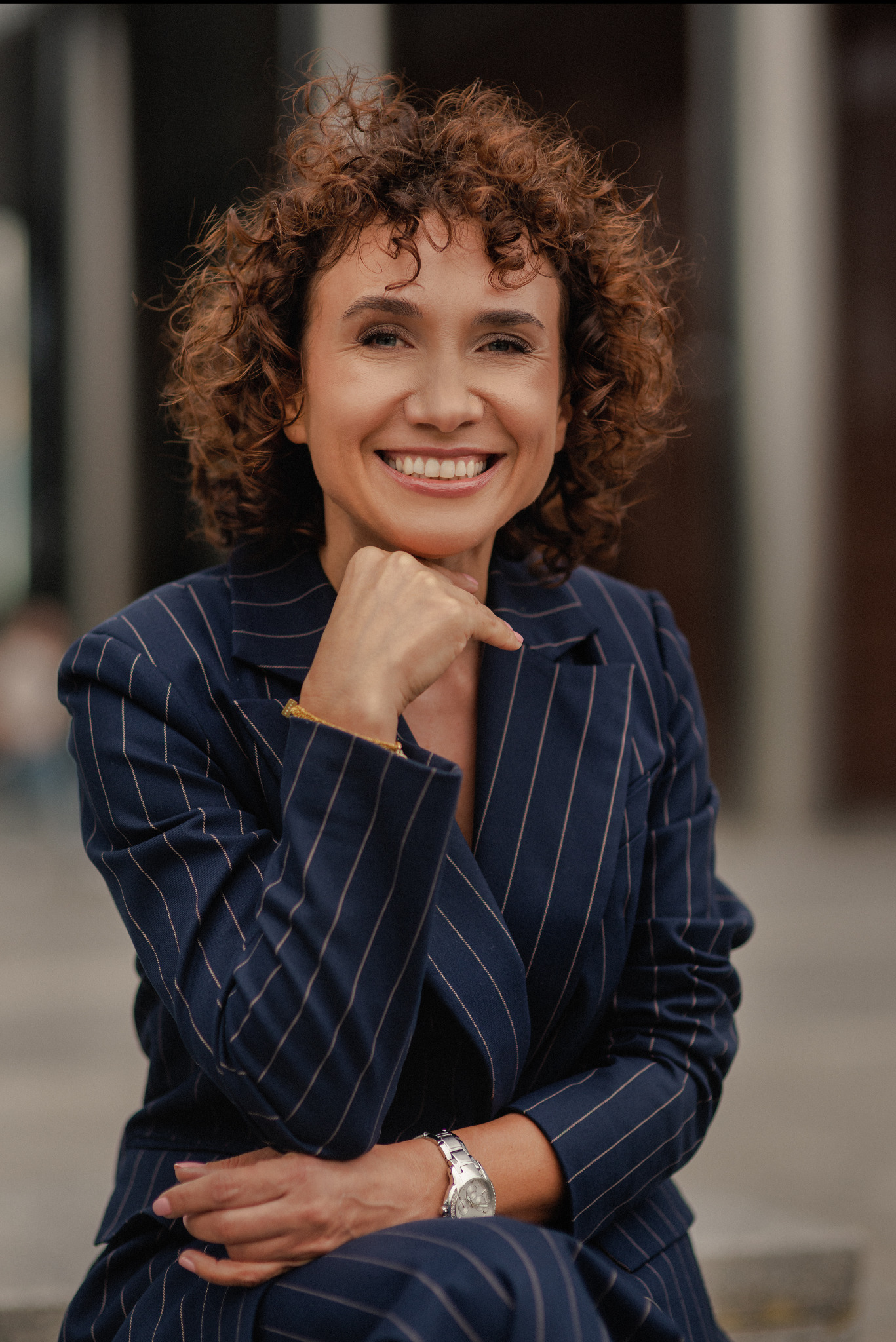 Uśmiechnięta kobieta w pinstripe garniturze, opierająca podbródek na dłoni, prezentuje profesjonalny wizerunek na neutralnym tle.