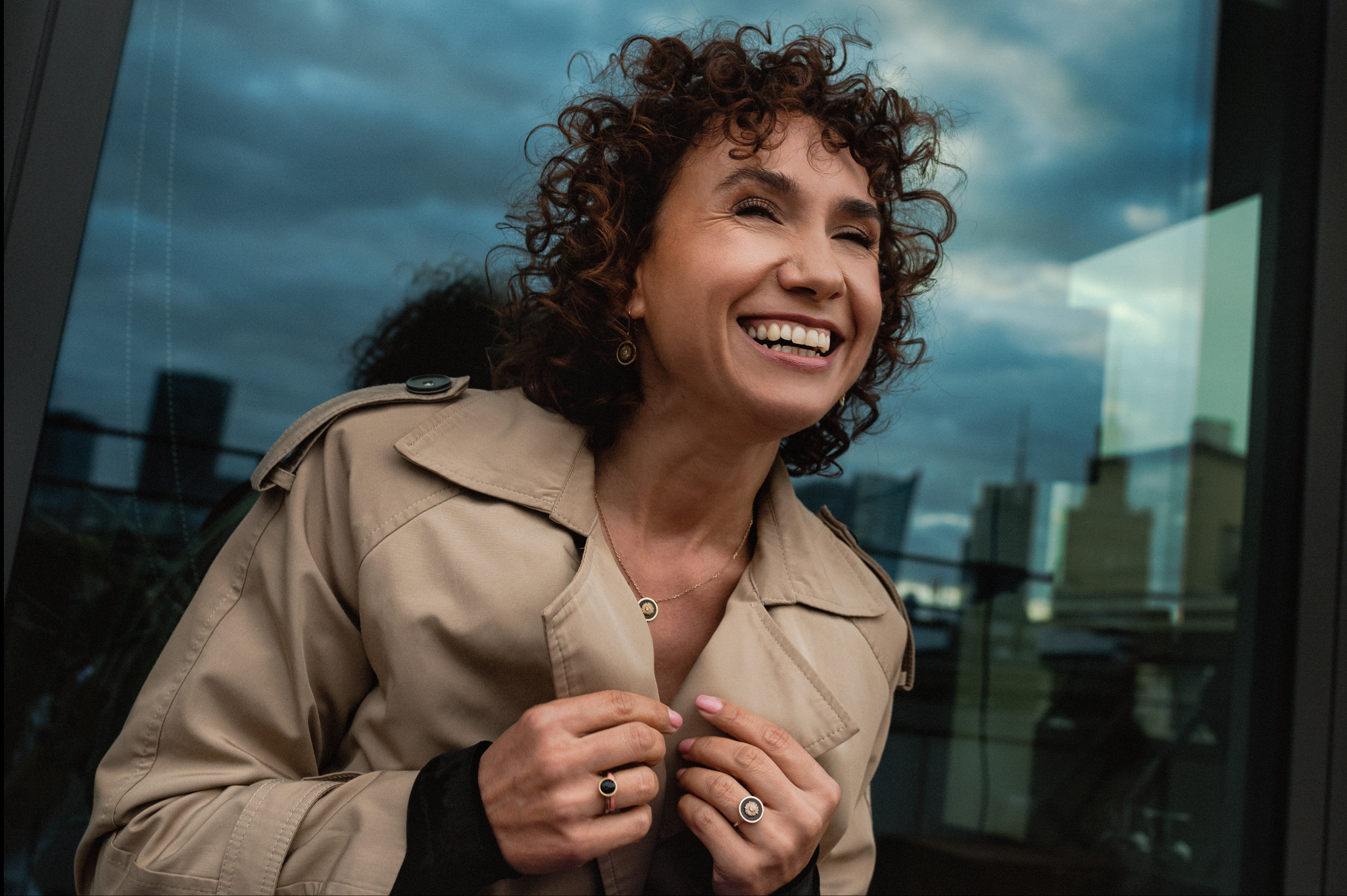 Uśmiechnięta kobieta w beżowym płaszczu, z kręconymi włosami i biżuterią, pozuje na tle szklanej fasady budynku odbijającej pochmurne niebo i miejski krajobraz.