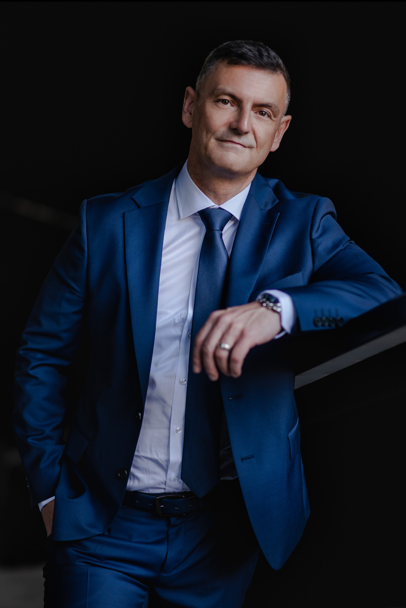 Mężczyzna w niebieskim garniturze opiera się o czarną barierkę, prezentując elegancki strój i zegarek. Uśmiech na twarzy dodaje profesjonalnego charakteru.