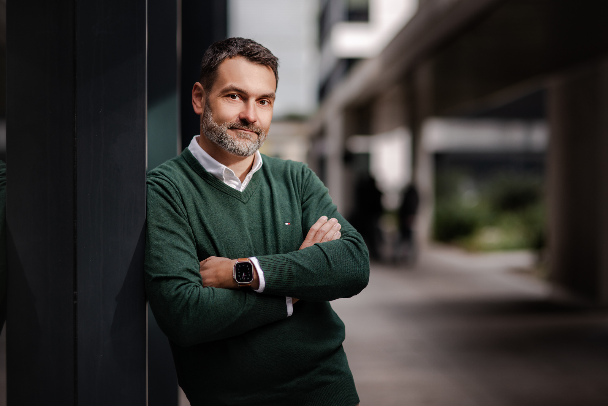 Mężczyzna z brodą w zielonym swetrze i białej koszuli, oparty o ciemny filar budynku z założonymi rękami, w tle rozmyty krajobraz miejski.