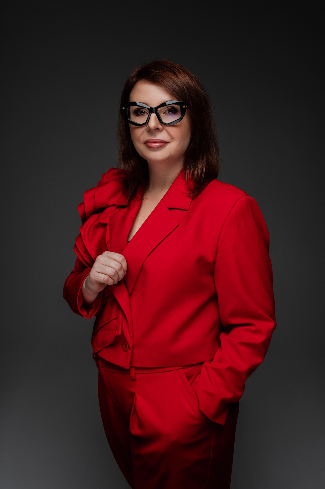 Portret kobiety w czerwonym garniturze i okularach na szarym tle. Profesjonalny wizerunek,  mocne oświetlenie,  wyraziste kolory,  odważna stylizacja.