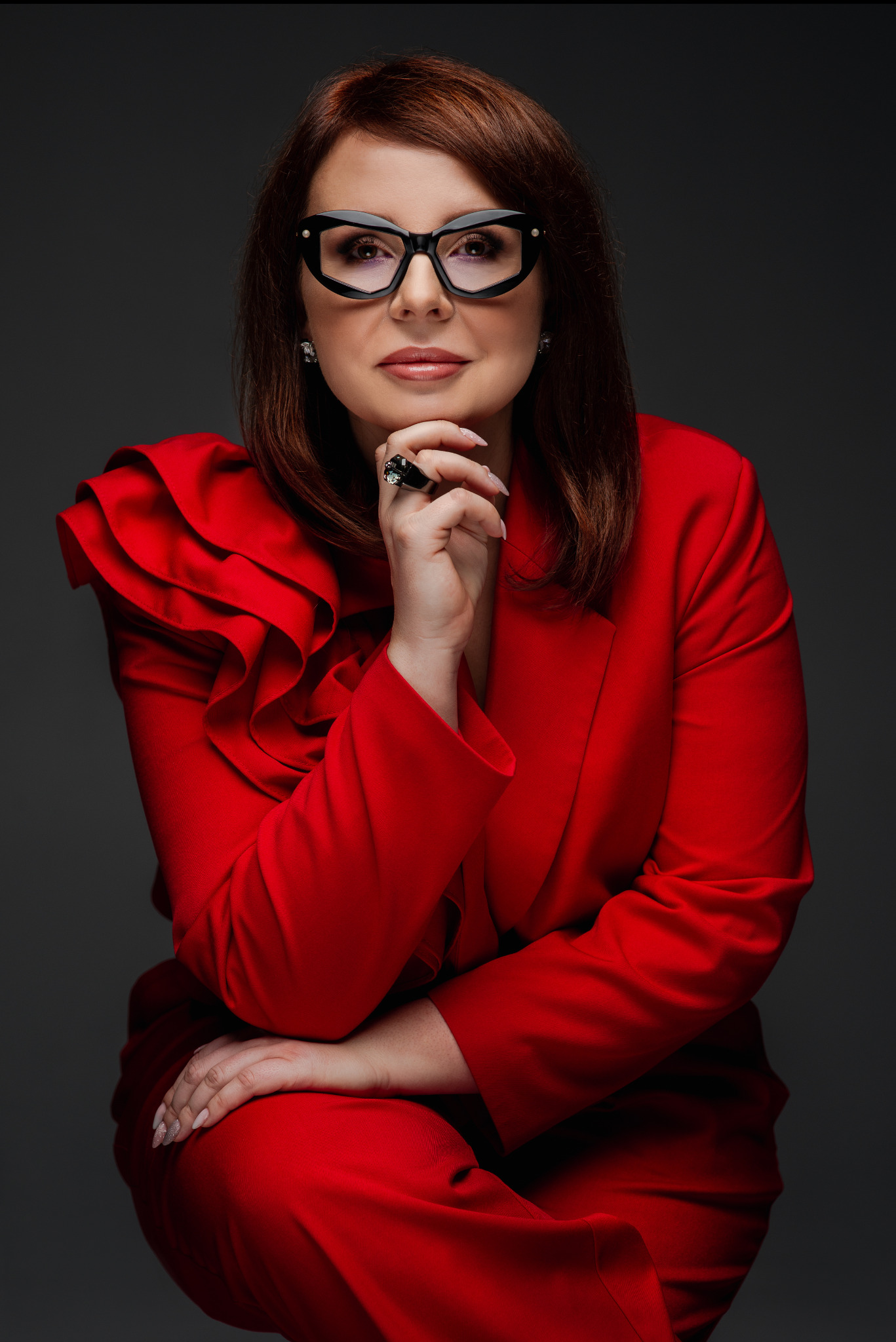 Portret kobiety w czerwonym kostiumie z falbanami, w okularach, z ręką pod brodą i dużym pierścieniem na palcu, na ciemnoszarym tle.