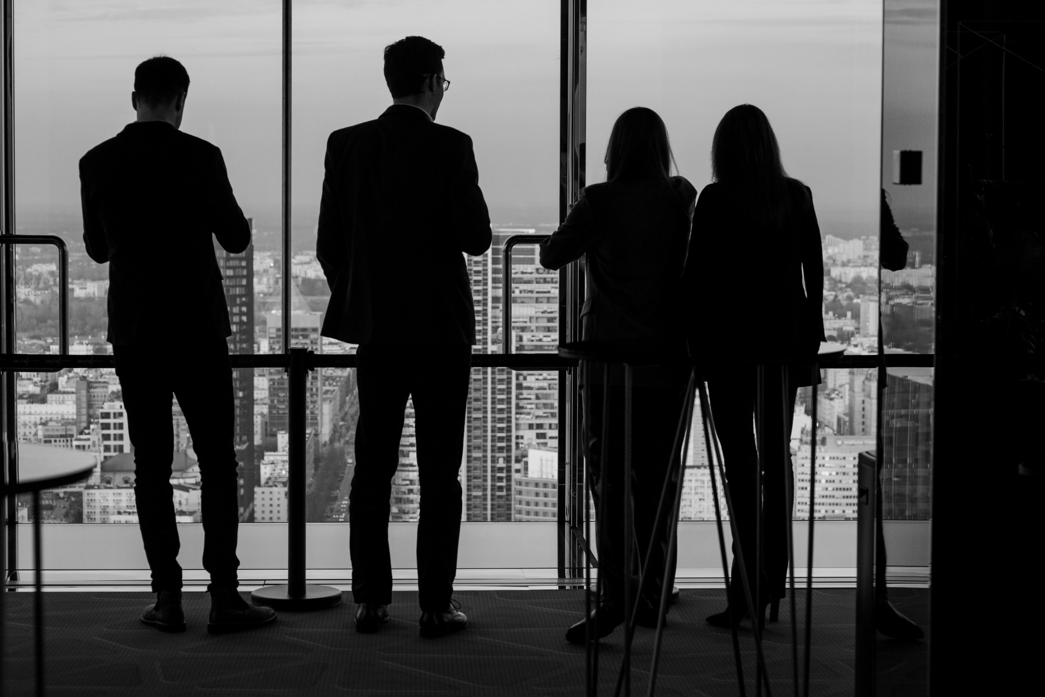 Czarno-białe zdjęcie sylwetek czterech osób w garniturach, stojących tyłem na tle okna z widokiem na miasto. Osoby wpatrują się w panoramę miejską.