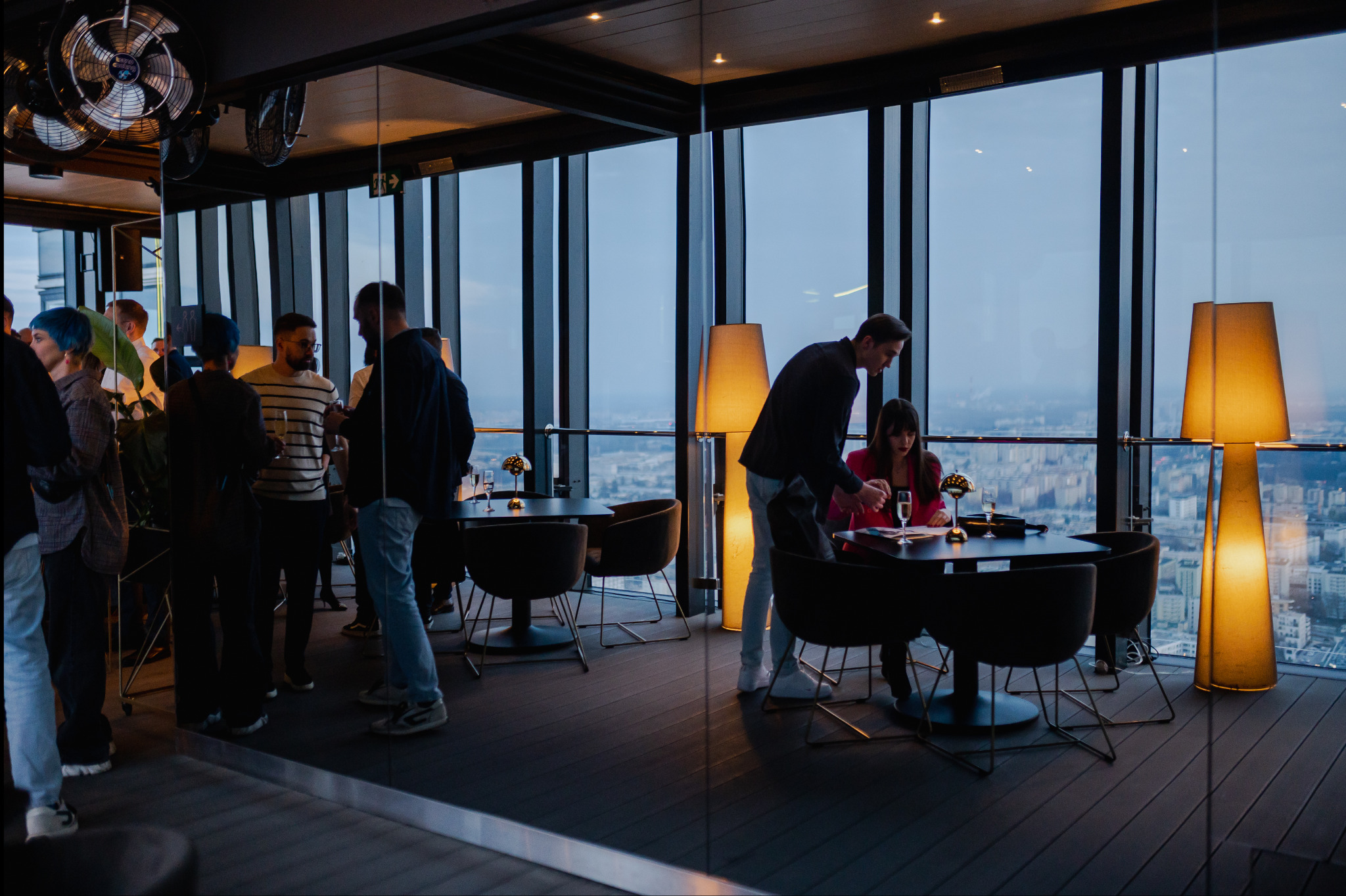 Event w nowoczesnym wnętrzu na wysokościach. Goście w swobodnych strojach, część przy stolikach, część w grupach. Duże okna z widokiem na miasto.