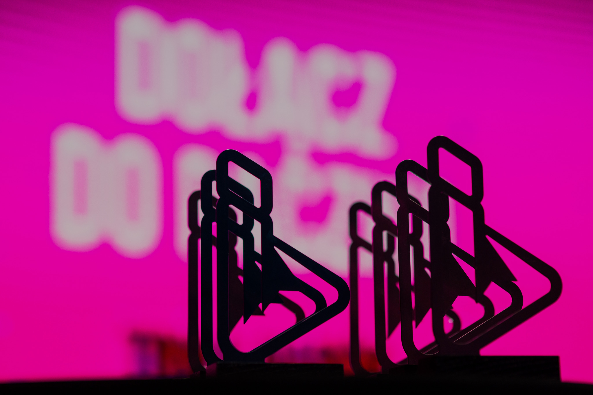 Rząd metalowych statuetek w kształcie kursora na tle różowego, rozmytym napisu 'Dołącz do...', prezentowany podczas gali eventowej.