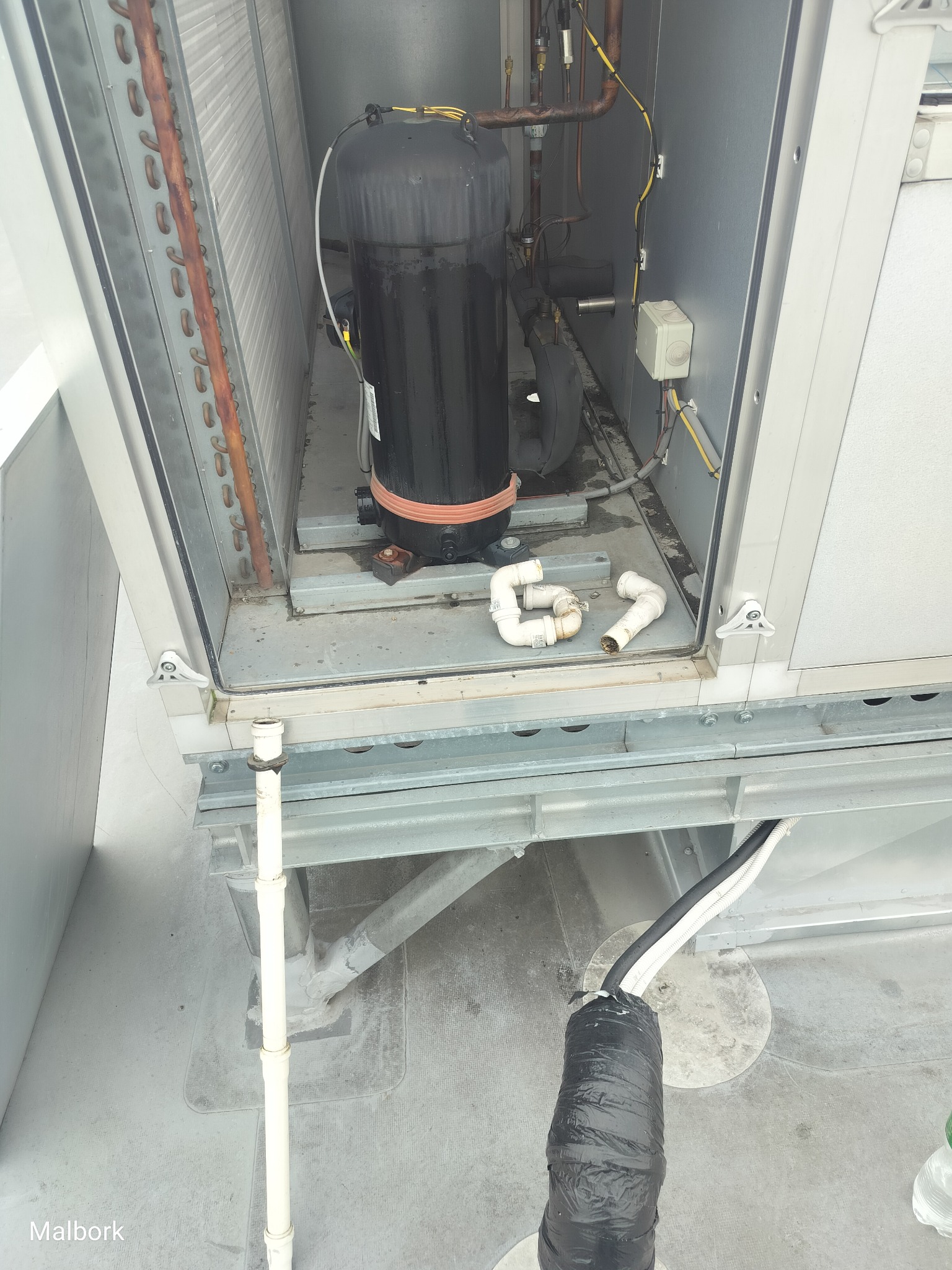 Otwarta obudowa urządzenia wentylacyjnego z widocznym kompresorem, rurami miedzianymi i elementami instalacji elektrycznej. Częściowo widoczne rury PCV i przewody.