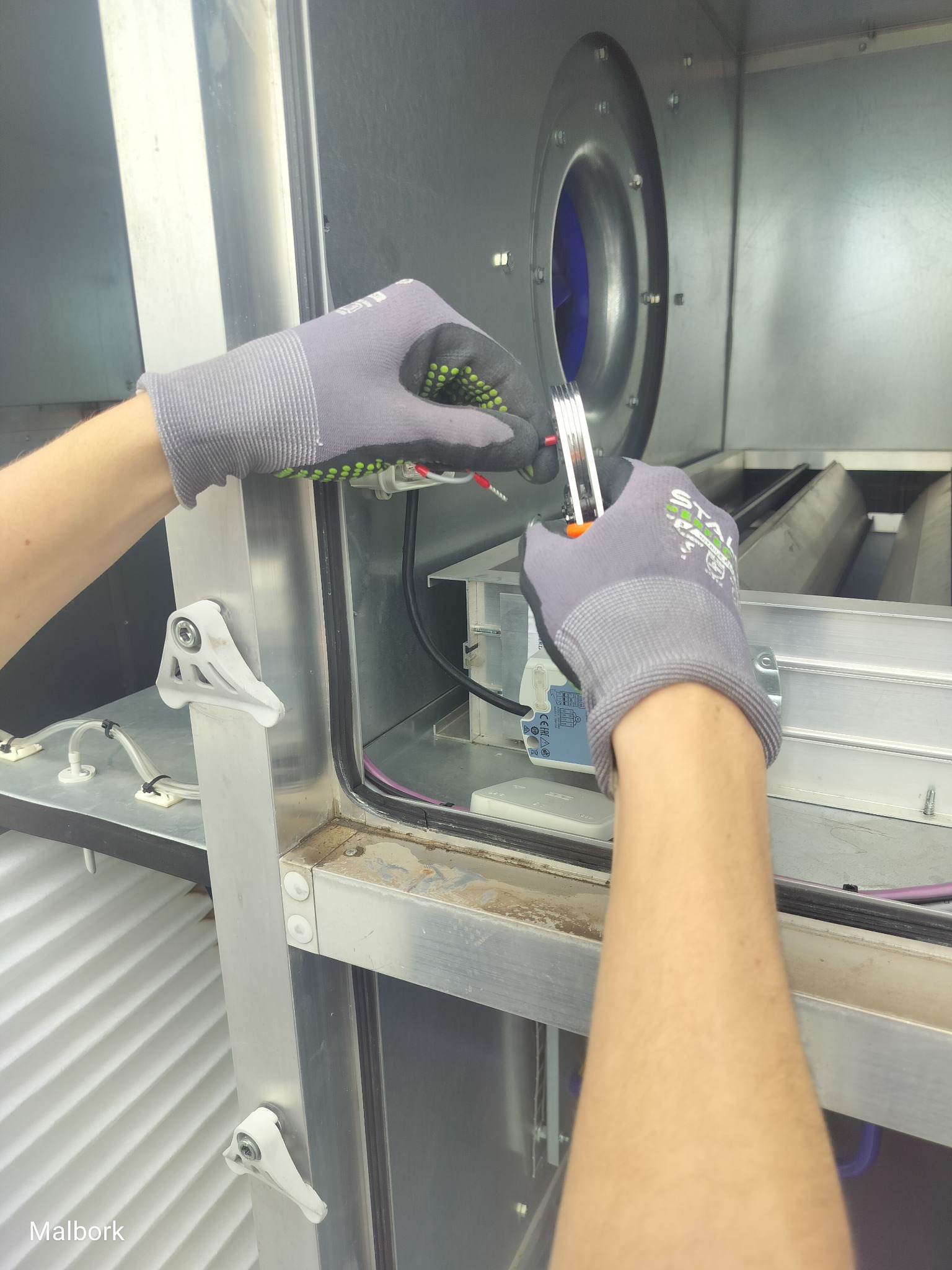 Prace montażowe wewnątrz metalowej obudowy wentylatora. Widoczne dłonie w rękawicach ochronnych, przewody i elementy instalacji. W tle wentylator.