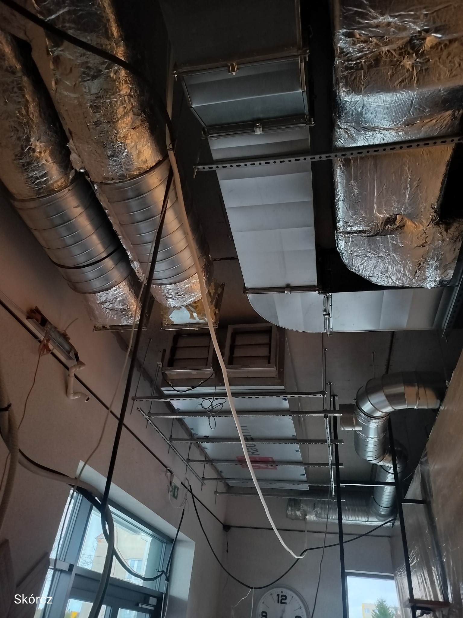 Widok na sufit z zamontowanymi przewodami wentylacyjnymi, rurami i elementami instalacji. Część instalacji jest w srebrnej izolacji. Widoczne okno i zegar.