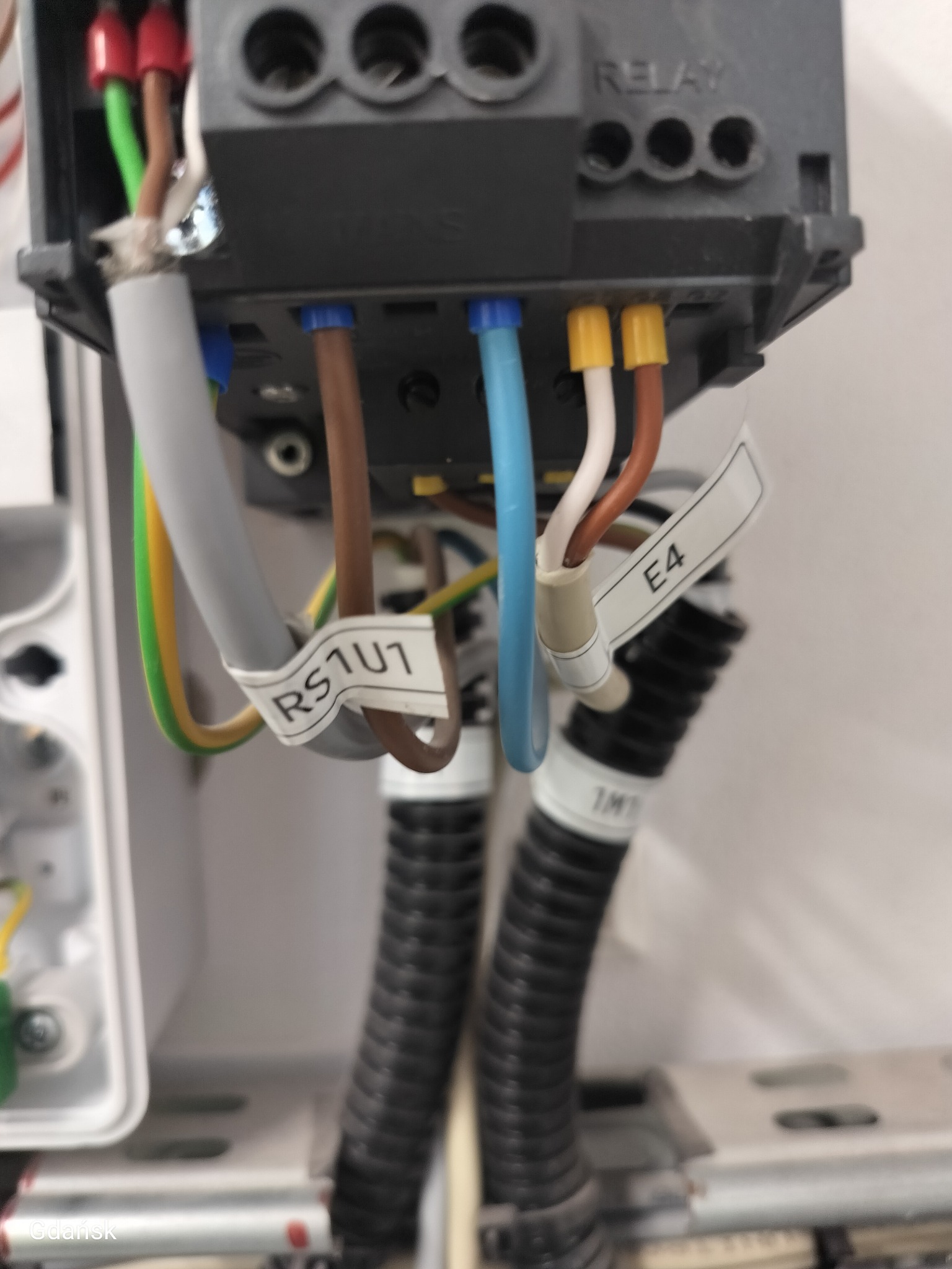 Szczegółowe ujęcie podłączenia przewodów o różnych kolorach (brązowy, niebieski, żółto-zielony) do listwy zaciskowej z oznaczeniami E4 i RS1U1.