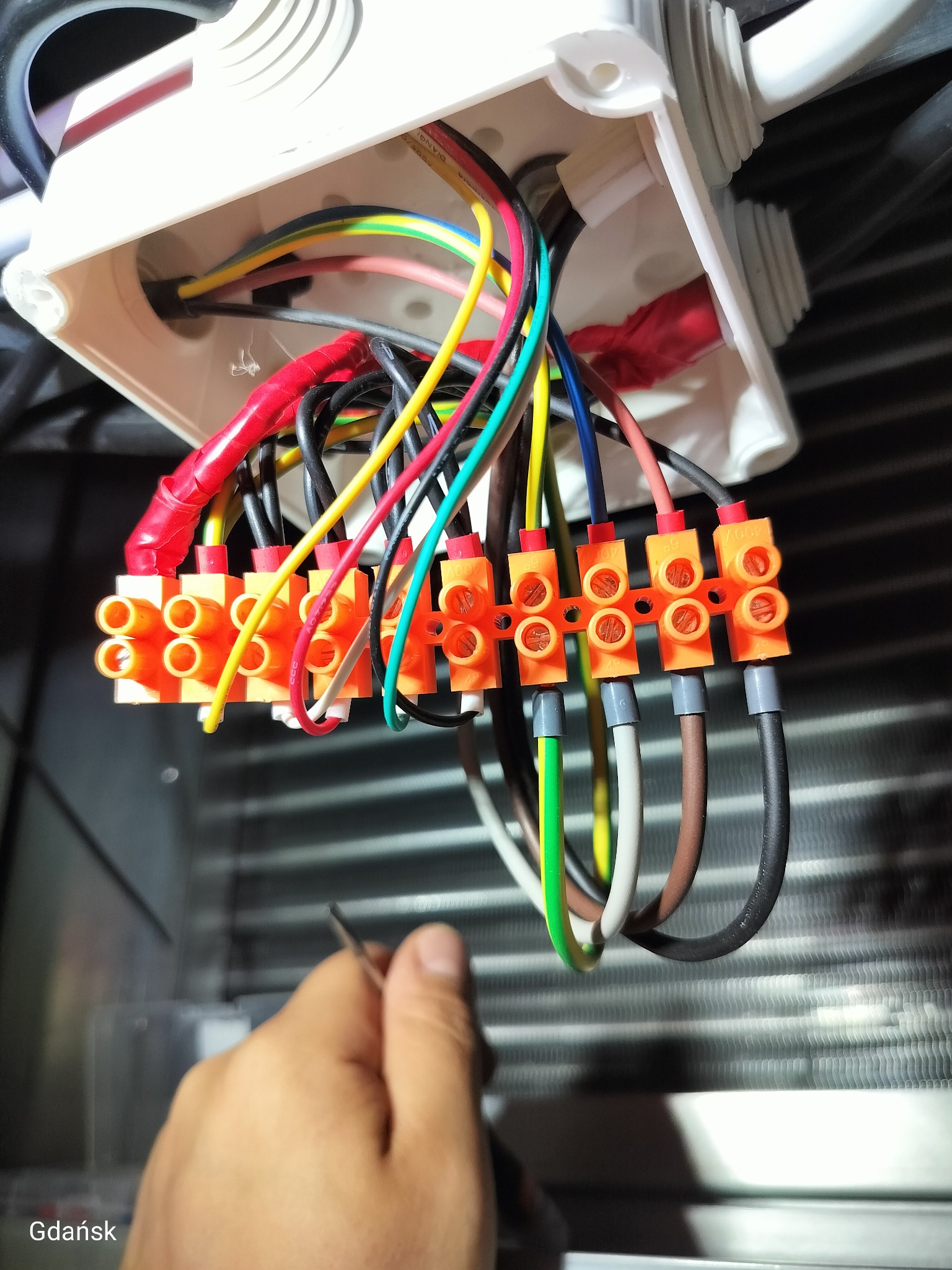Rozdzielnica elektryczna z podłączonymi przewodami o różnych kolorach, widoczne złączki Wago i fragment ręki elektryka z narzędziem w tle chłodnicy.