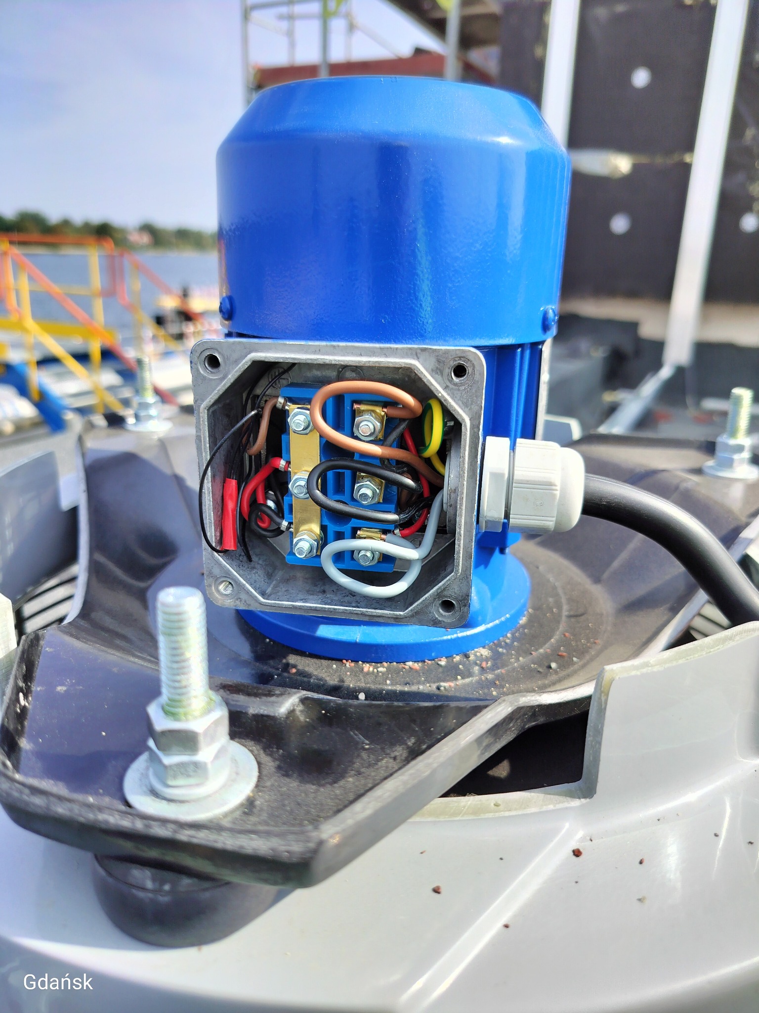 Niebieski silnik elektryczny z otwartą skrzynką przyłączeniową, widoczne okablowanie i złącza. Montaż na szarej podstawie, tło z elementami krajobrazu.