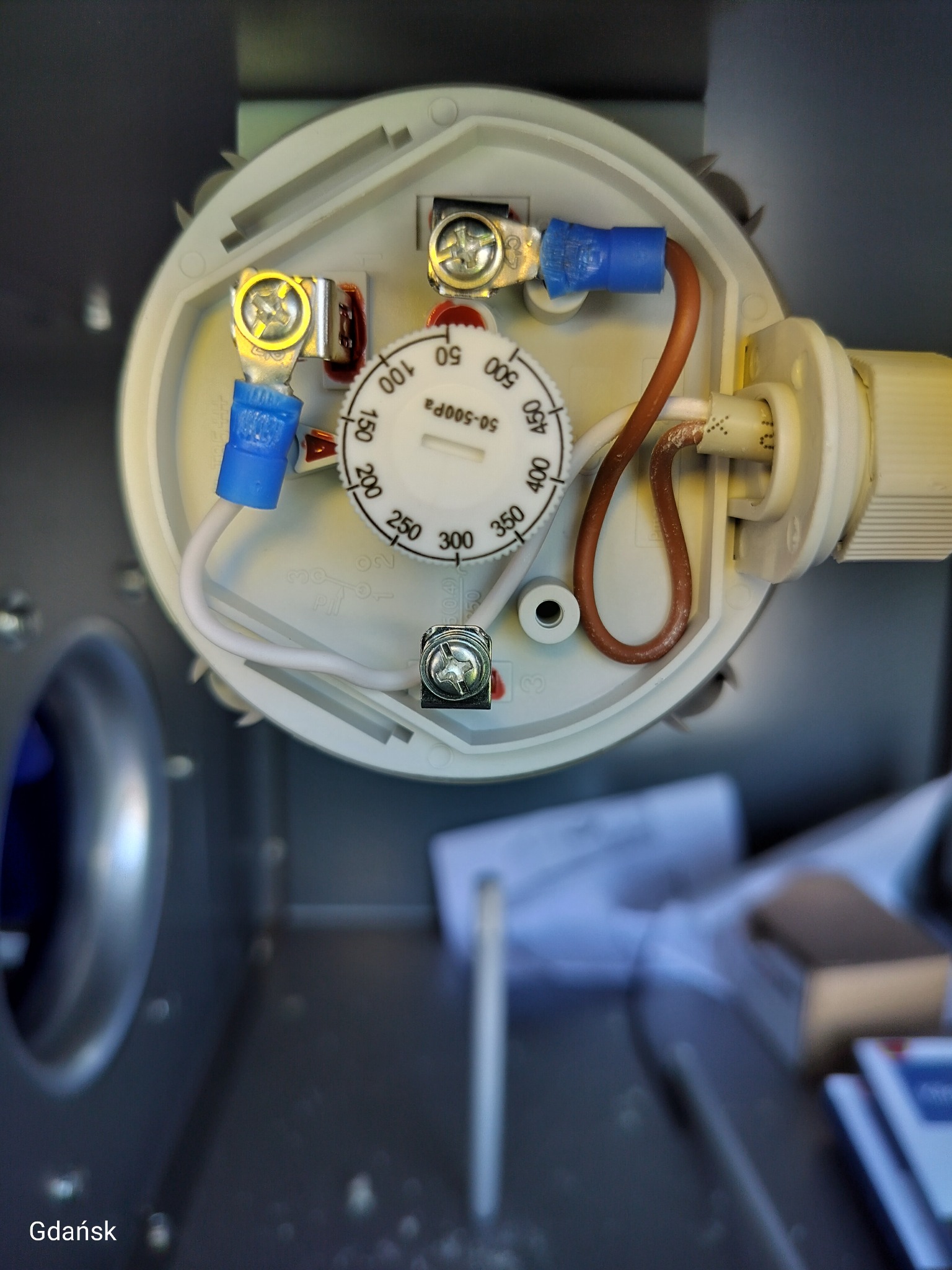 Wnętrze okrągłego regulatora ciśnienia w instalacji wentylacyjnej z widocznymi przewodami i ustawieniami skali. Widok z góry na podzespoły urządzenia.