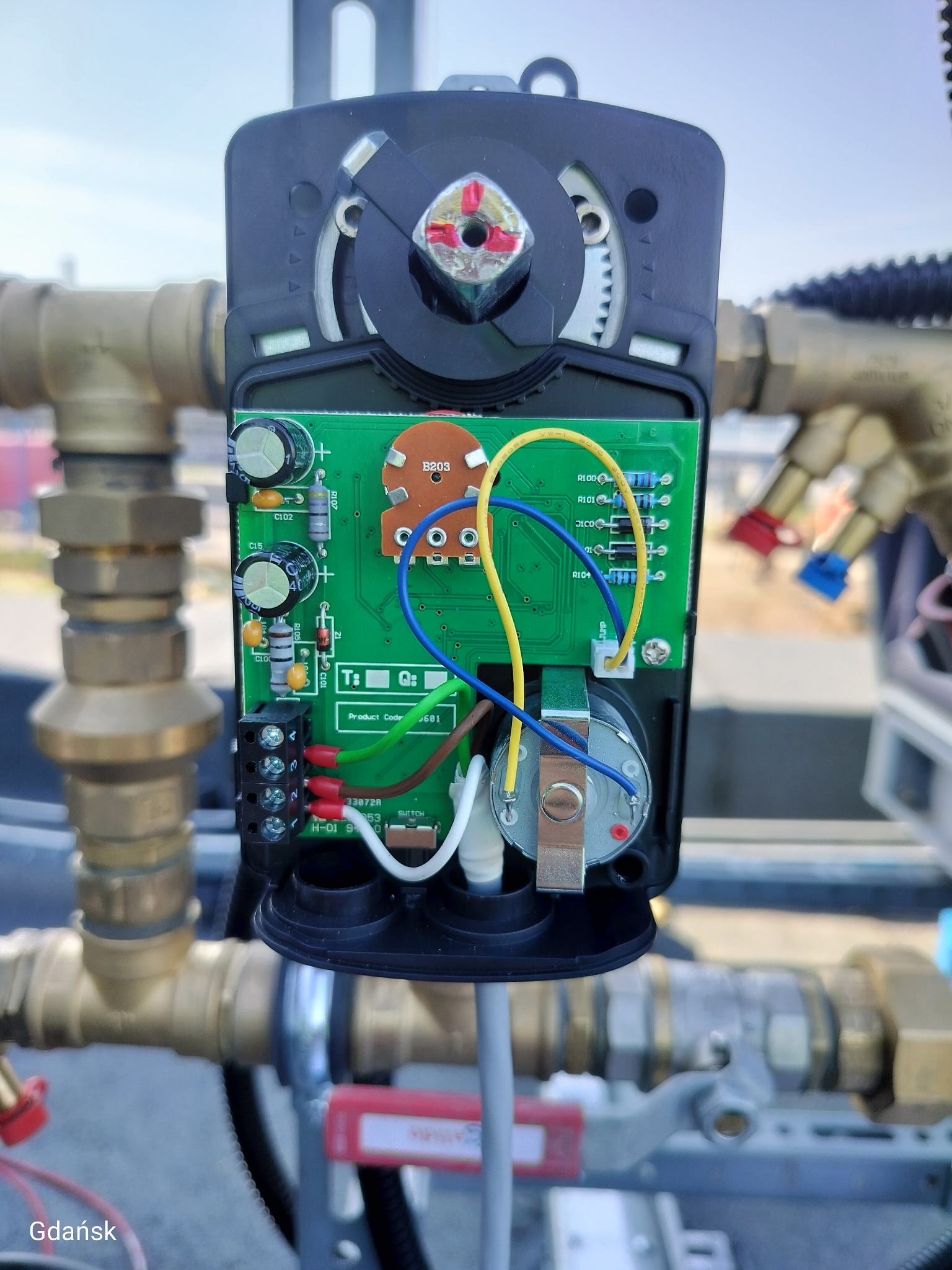 Szczegółowe ujęcie zaworu z odsłoniętą elektroniką, widoczne przewody, kondensatory i elementy instalacji hydraulicznej w tle. Dominuje zieleń płytki i złoto rur.