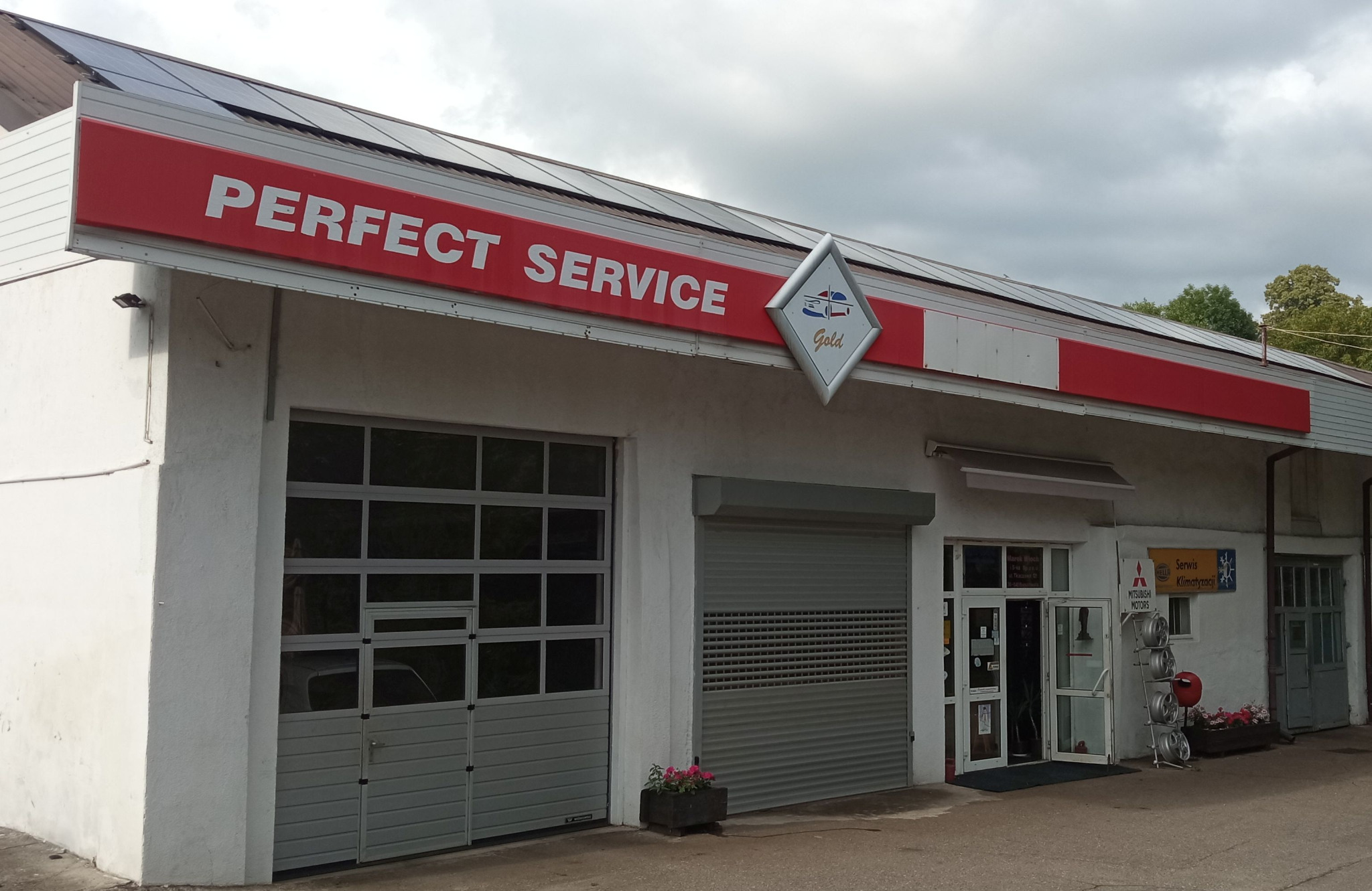 Fasada budynku warsztatu samochodowego 'Perfect Service Gold' z widocznymi bramami wjazdowymi i logo firmy. Na dachu panele solarne.