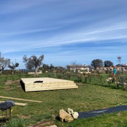 Oleh Tuz EUROBILD - Drewniany podest tarasu w trakcie budowy na zielonej trawie, z elementami narzędzi i roślinności w tle, pod błękitnym niebem. Widoczne deski i elementy konstrukcyjne.