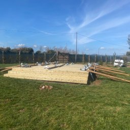 Oleh Tuz EUROBILD - Drewniany taras w trakcie budowy z elementami konstrukcyjnymi i metalowymi łącznikami na trawniku, w tle ogrodzenie i drzewa pod błękitnym niebem.