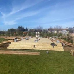 Budowa tarasu z drewna na trawie w słoneczny dzień. Widoczne deski, belki i elementy montażowe. Częściowo ukończona konstrukcja z widocznym tłem ogrodu.