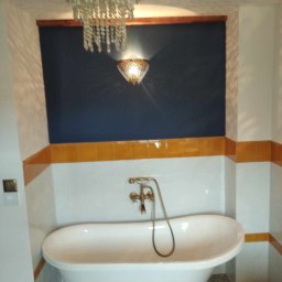 Oleh Tuz EUROBILD - Biała wanna na złotych nóżkach w łazience z ciemnoniebieską ścianą, oświetlona ozdobnym żyrandolem i kinkietem. Żółte płytki stanowią akcent kolorystyczny.